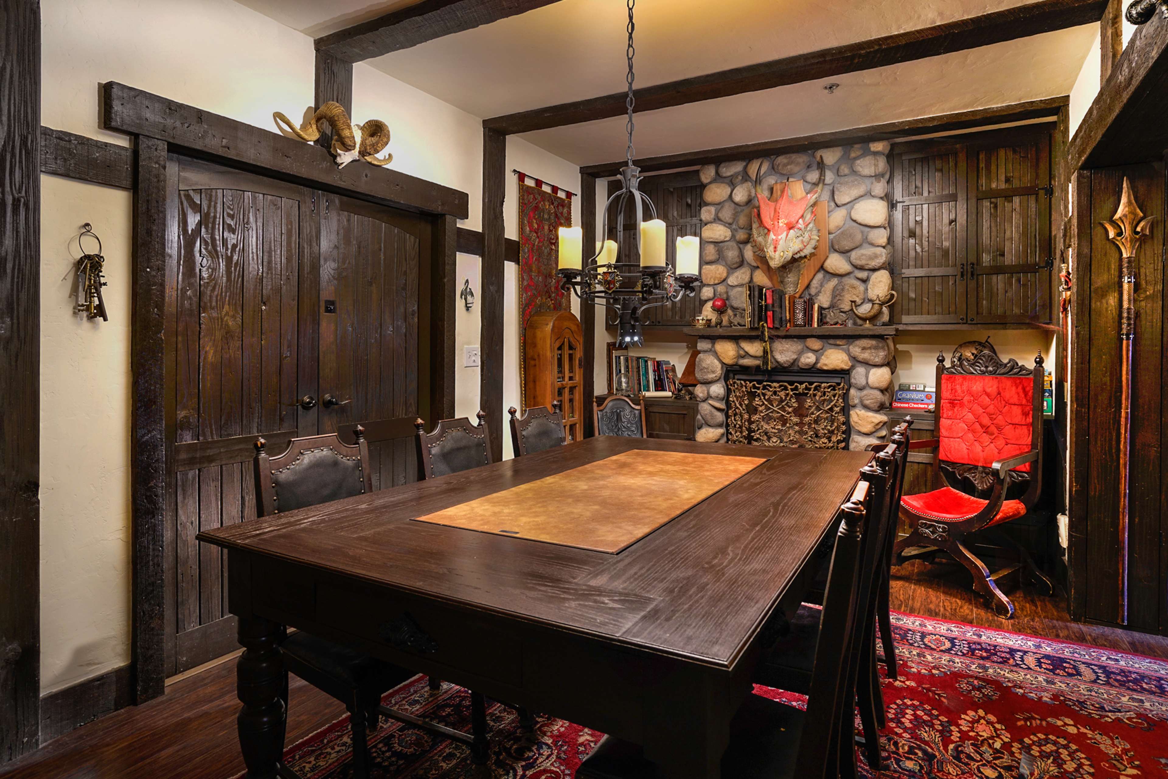 A rustic dining room features a large wooden table, surrounded by ornate chairs, with a stone fireplace and a mounted animal head decoratively positioned on the wall.