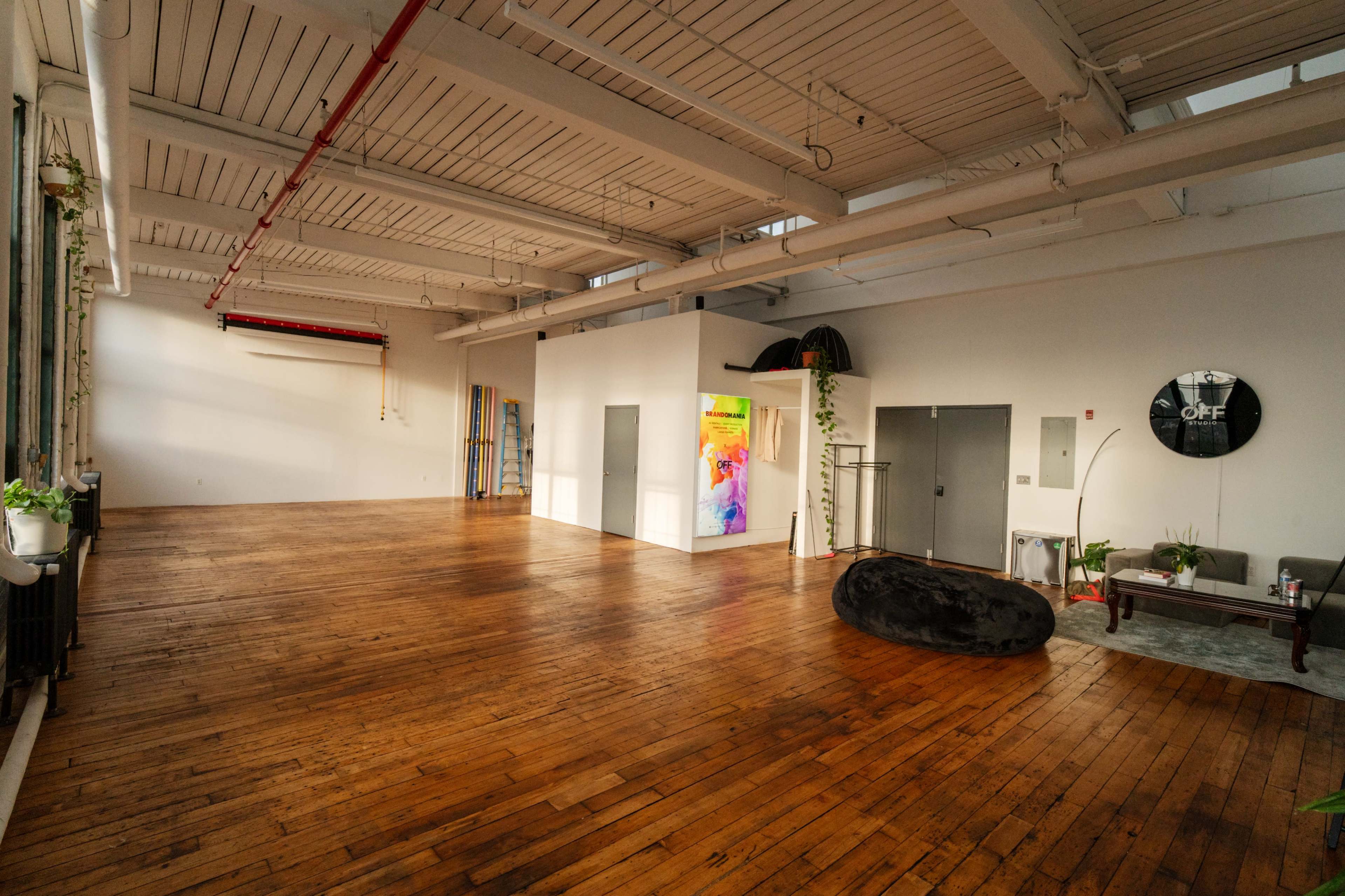 The image shows a spacious, empty interior with wooden flooring, large windows, and a few pieces of furniture and decor.