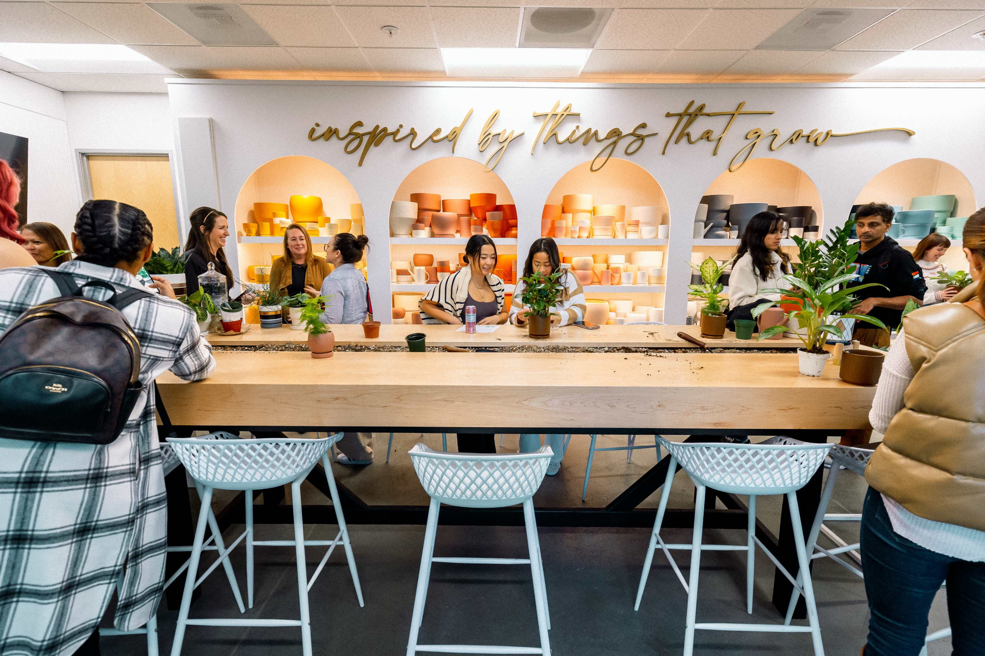 A group of people gather around a long table in a bright space filled with plants and decorative pottery, engaged in a hands-on planting activity.