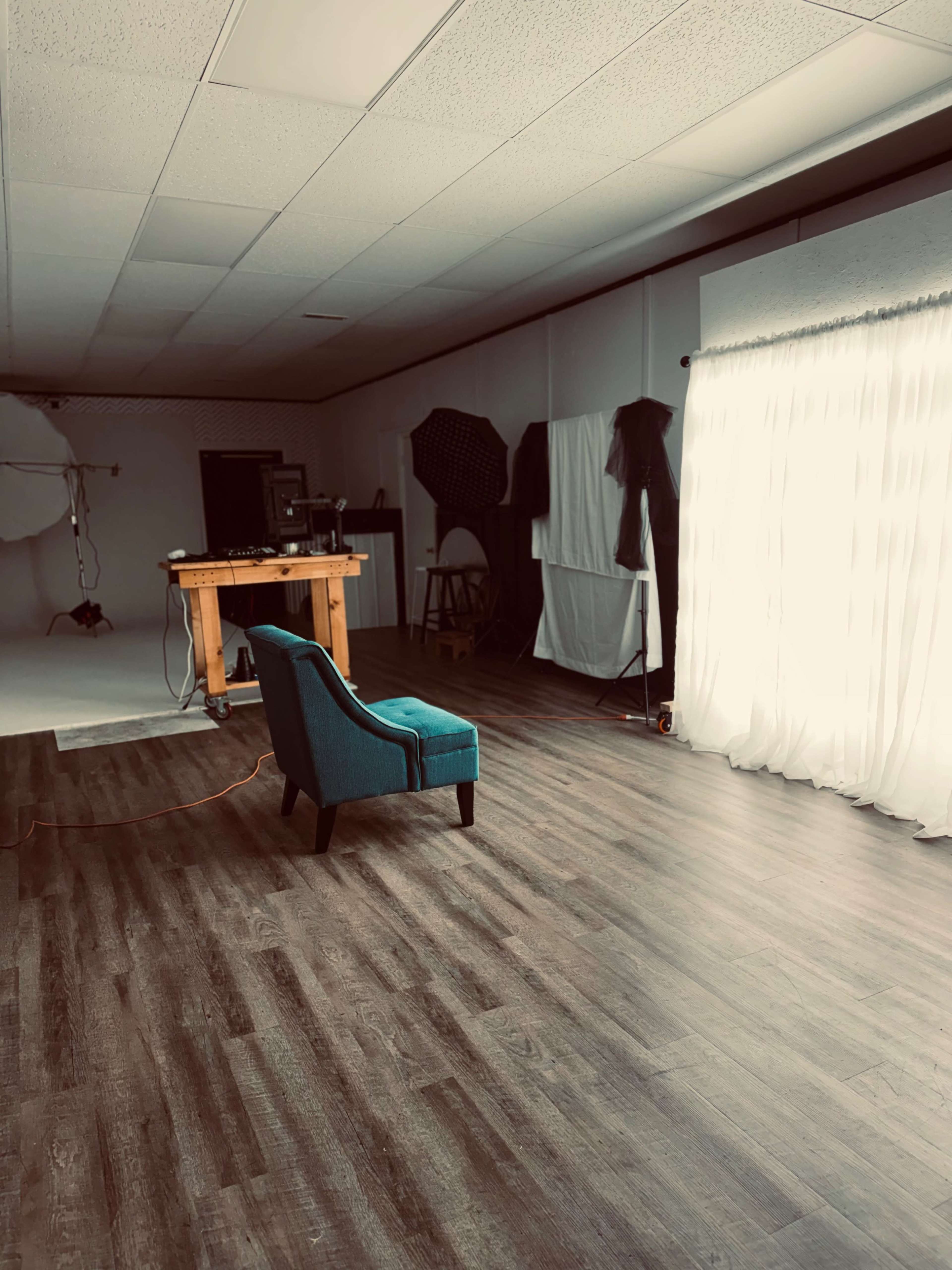 The image shows a spacious photography studio with a wooden table, a teal chair, and soft white curtains letting in natural light.