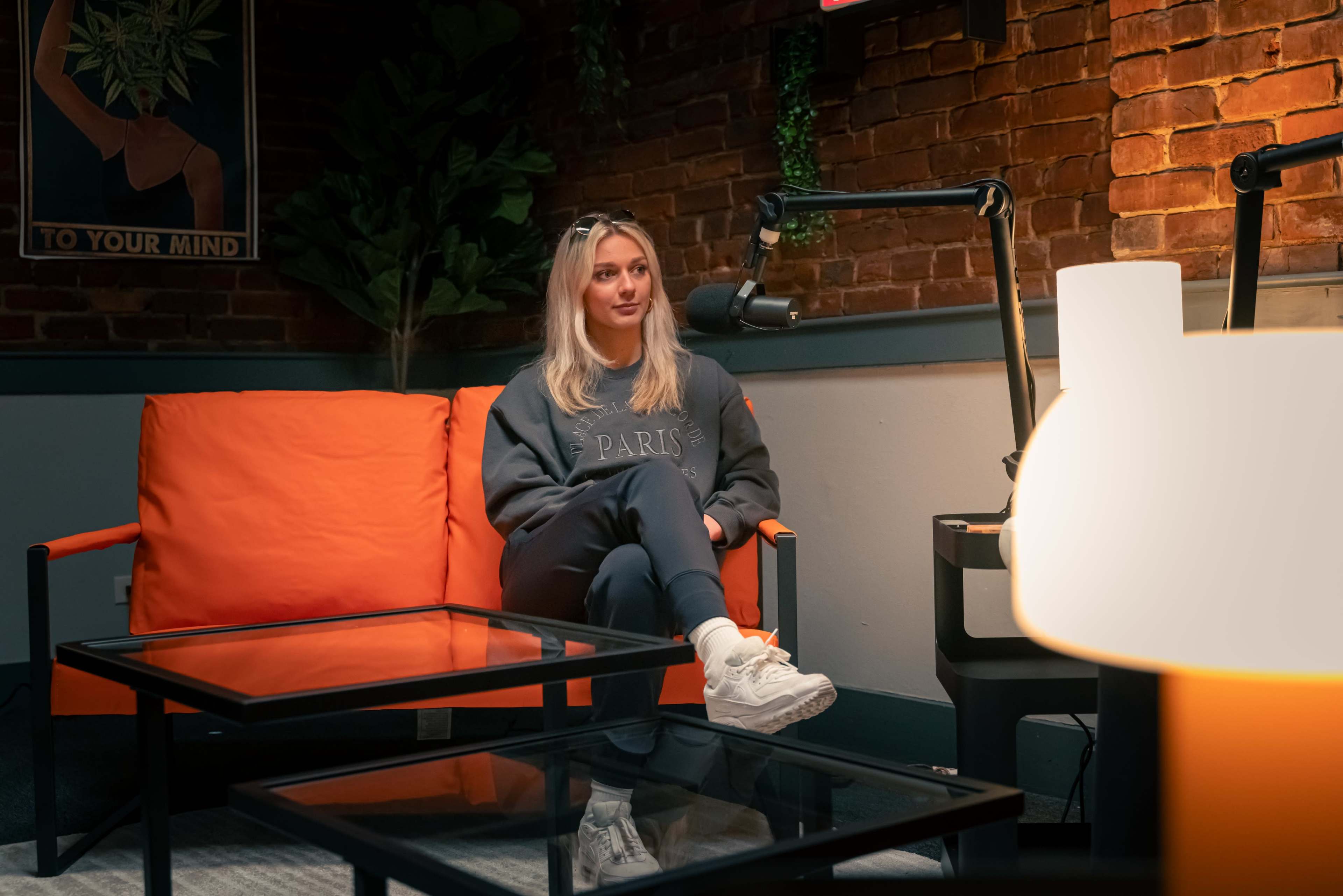 A person sits on an orange couch in a cozy room with exposed brick walls and a microphone setup nearby.