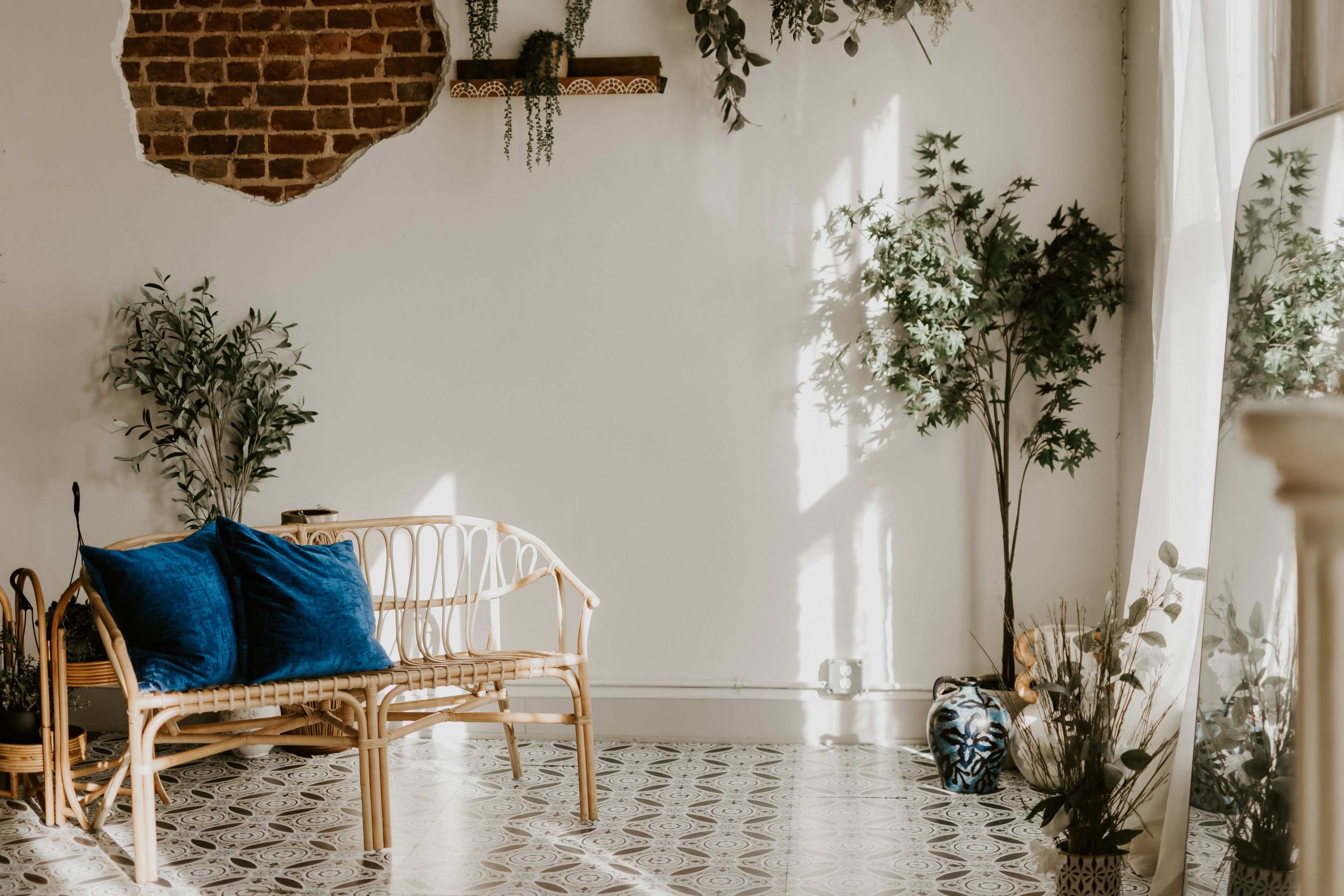 The image shows a bright, airy room featuring a rattan bench with blue pillows, indoor plants, and a patterned floor.