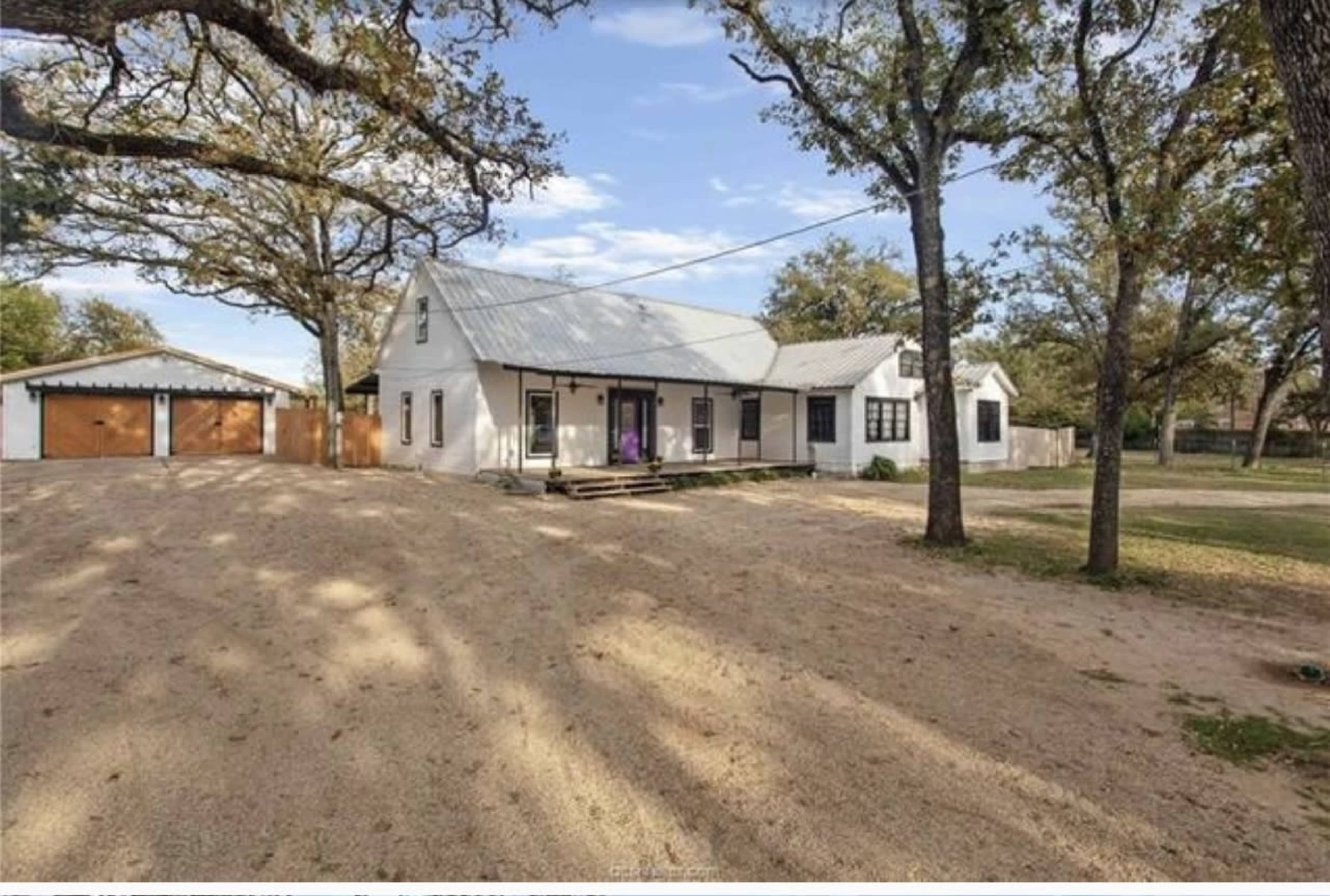 The image shows a white house with a metal roof, surrounded by gravel and trees, with a shed visible in the background.