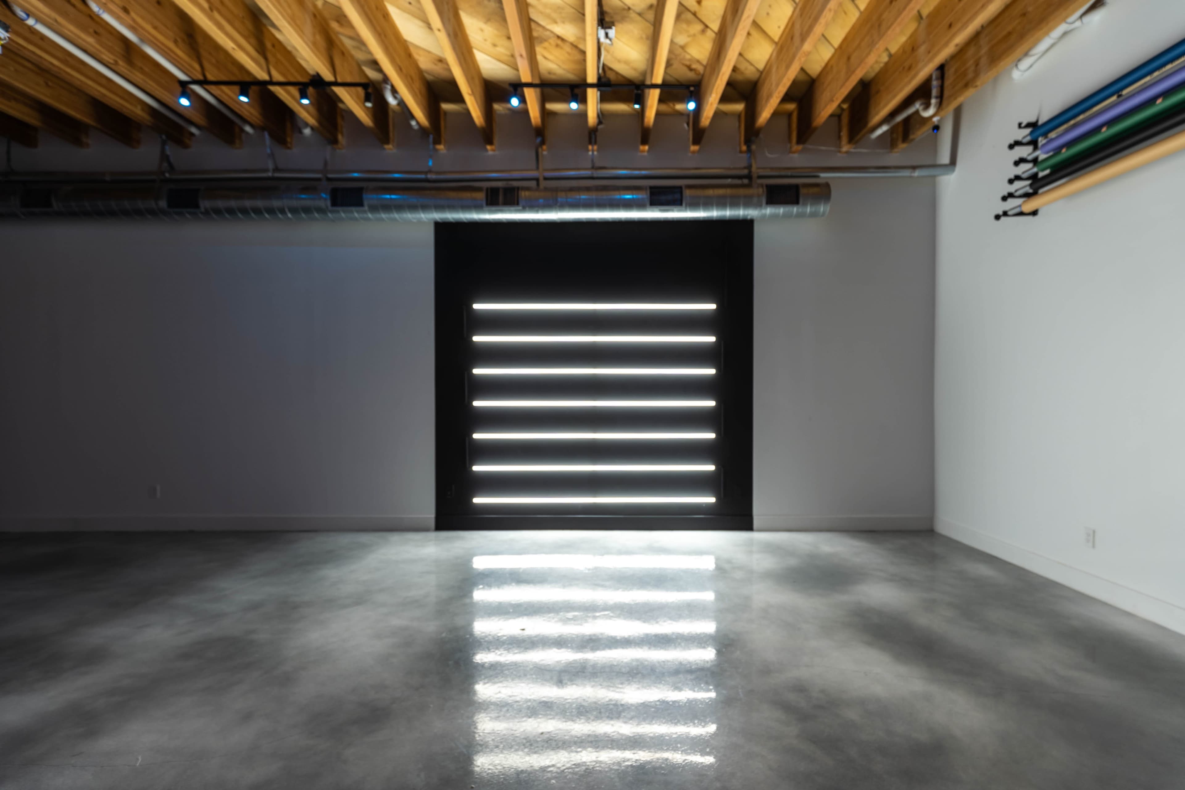 A spacious room with a concrete floor features a series of horizontal light strips on a dark wall, under a wooden ceiling with exposed beams.