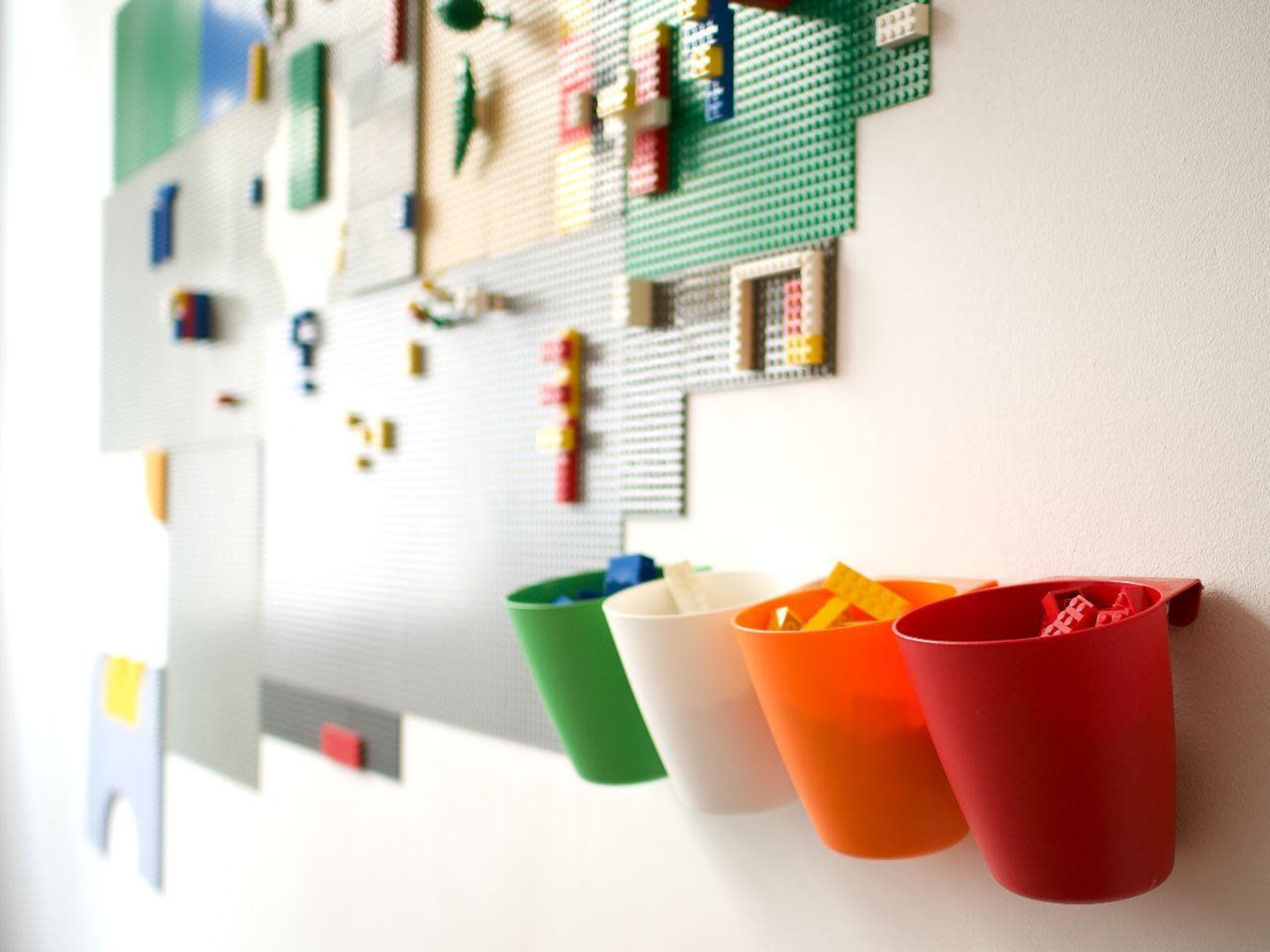 A white wall features a colorful LEGO building area with four hanging containers in green, white, orange, and red, each filled with assorted LEGO pieces.