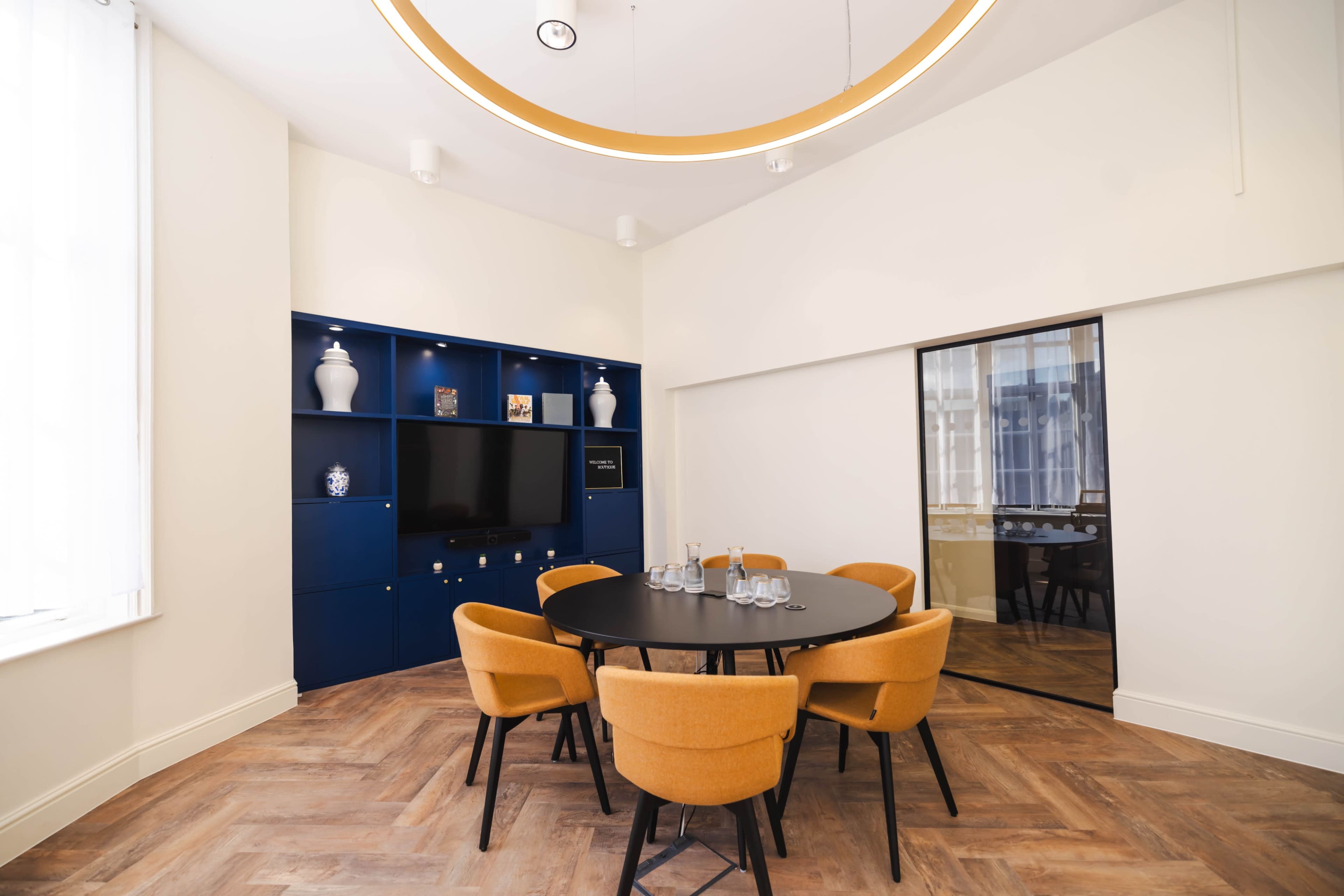A meeting room features a circular black table surrounded by orange chairs, with a blue shelving unit and a large mirror on the wall.