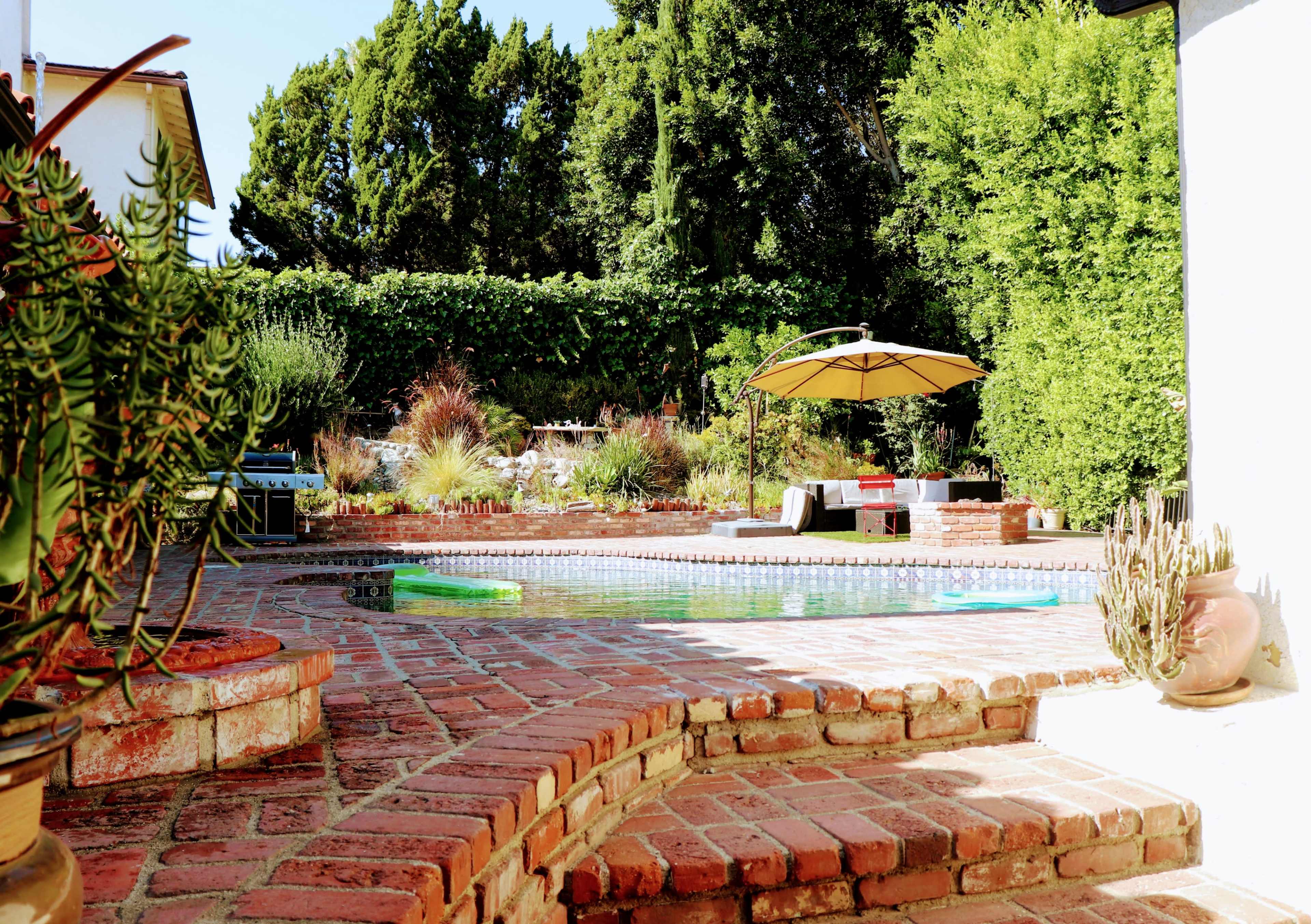 A landscaped backyard featuring a swimming pool surrounded by brick paving, lounge furniture, and an umbrella.