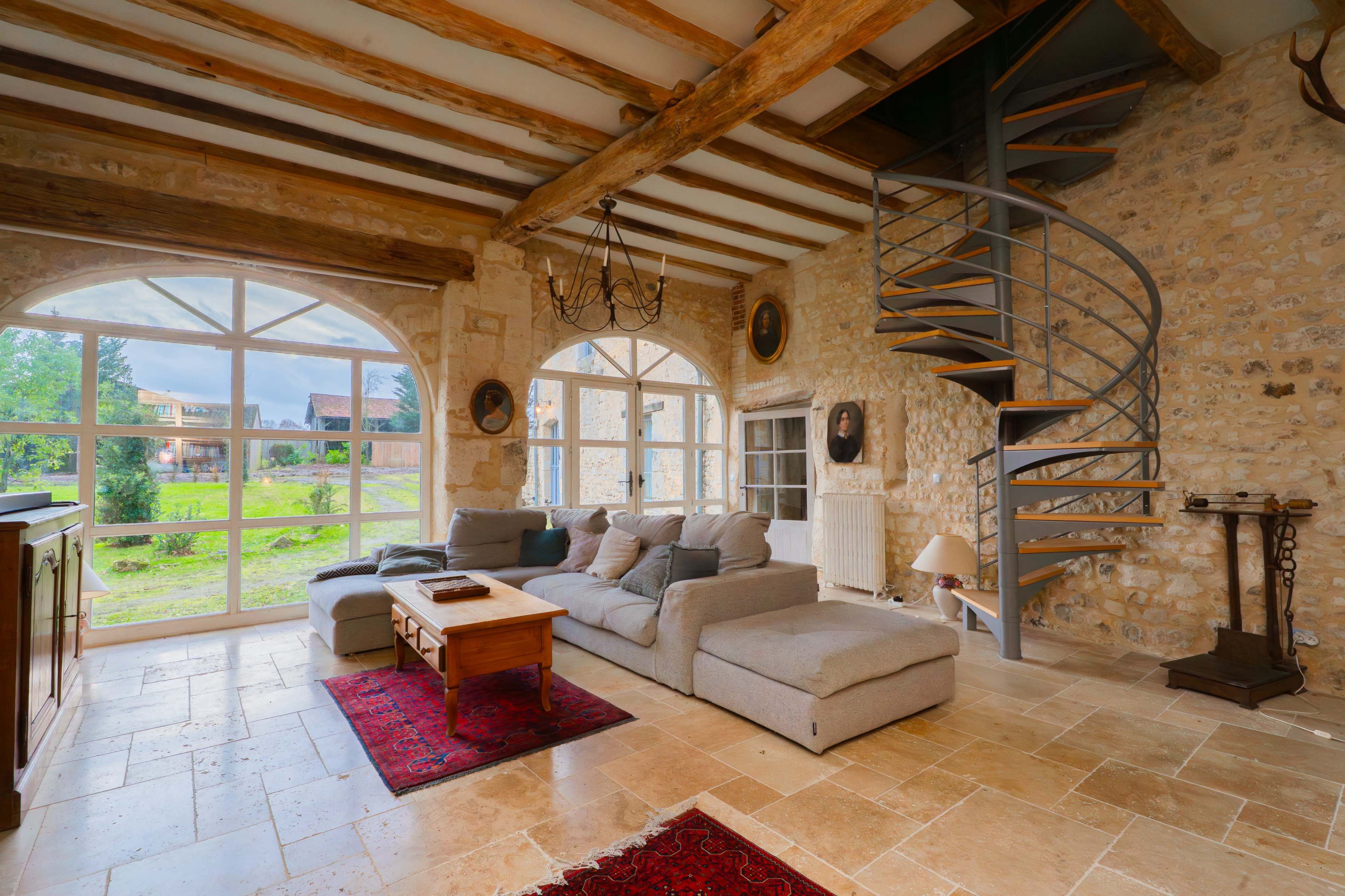 The image shows a spacious living room with a circular staircase, large arched windows, and stone walls, furnished with a light gray sectional sofa and a wooden coffee table.