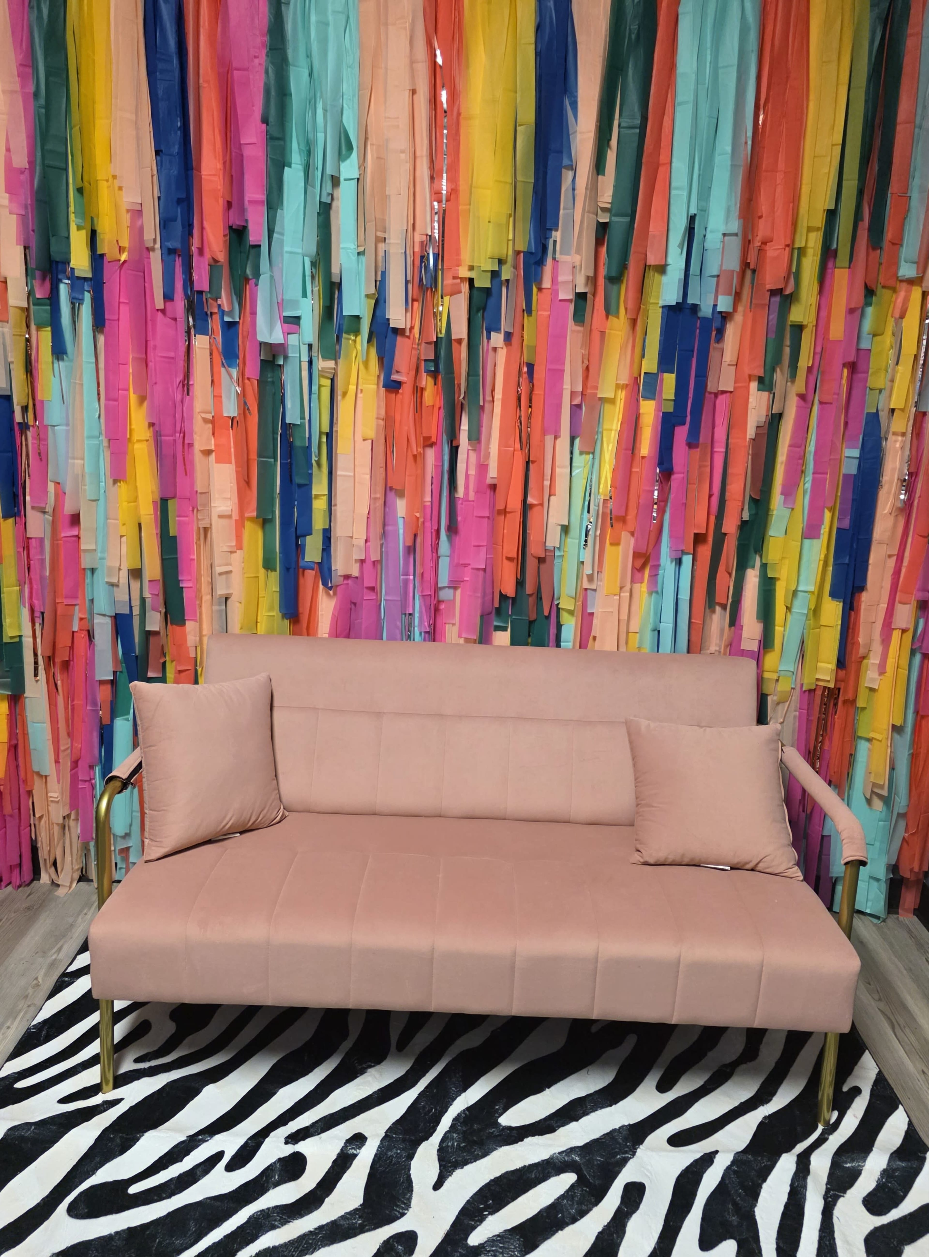 A pink sofa with two cushions is positioned in front of a vibrant, multi-colored backdrop of fabric strips, set on a zebra-print rug.