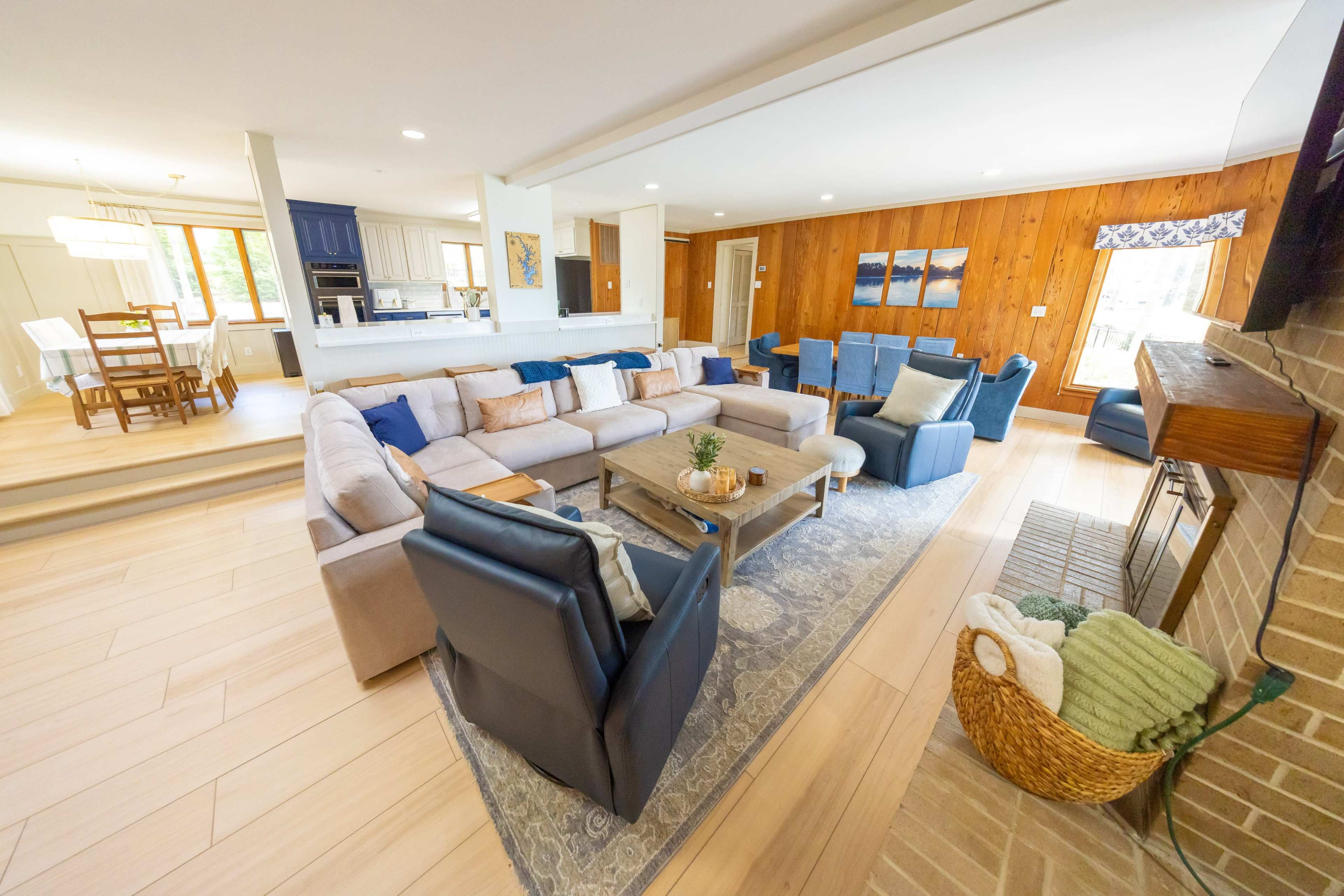 The image shows a spacious living room featuring a large sectional sofa, multiple armchairs, a coffee table, and a fireplace, with light-colored wood flooring and natural light coming through the windows.