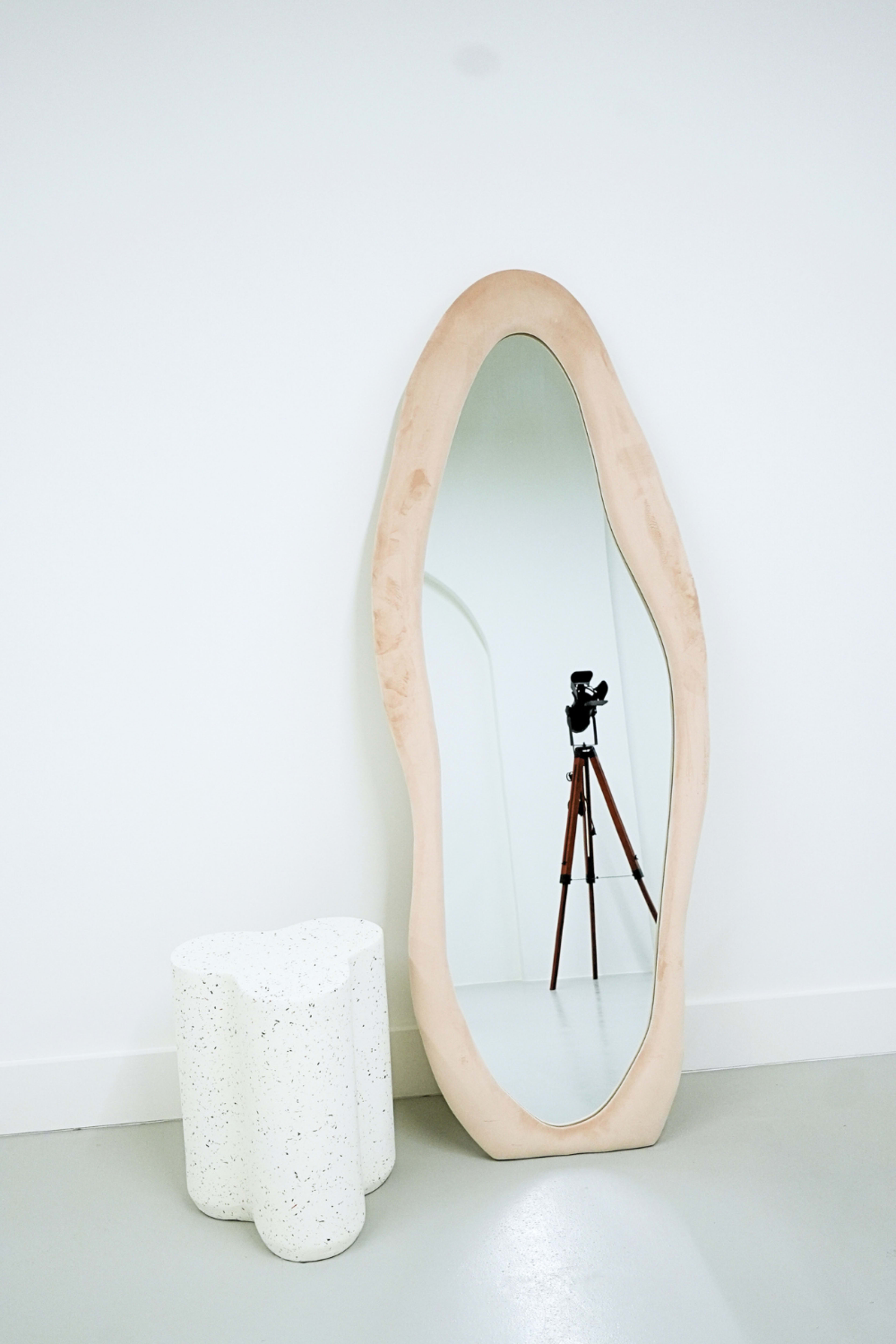 A large, irregularly shaped mirror with a soft pink frame leans against a white wall beside a textured white stool.