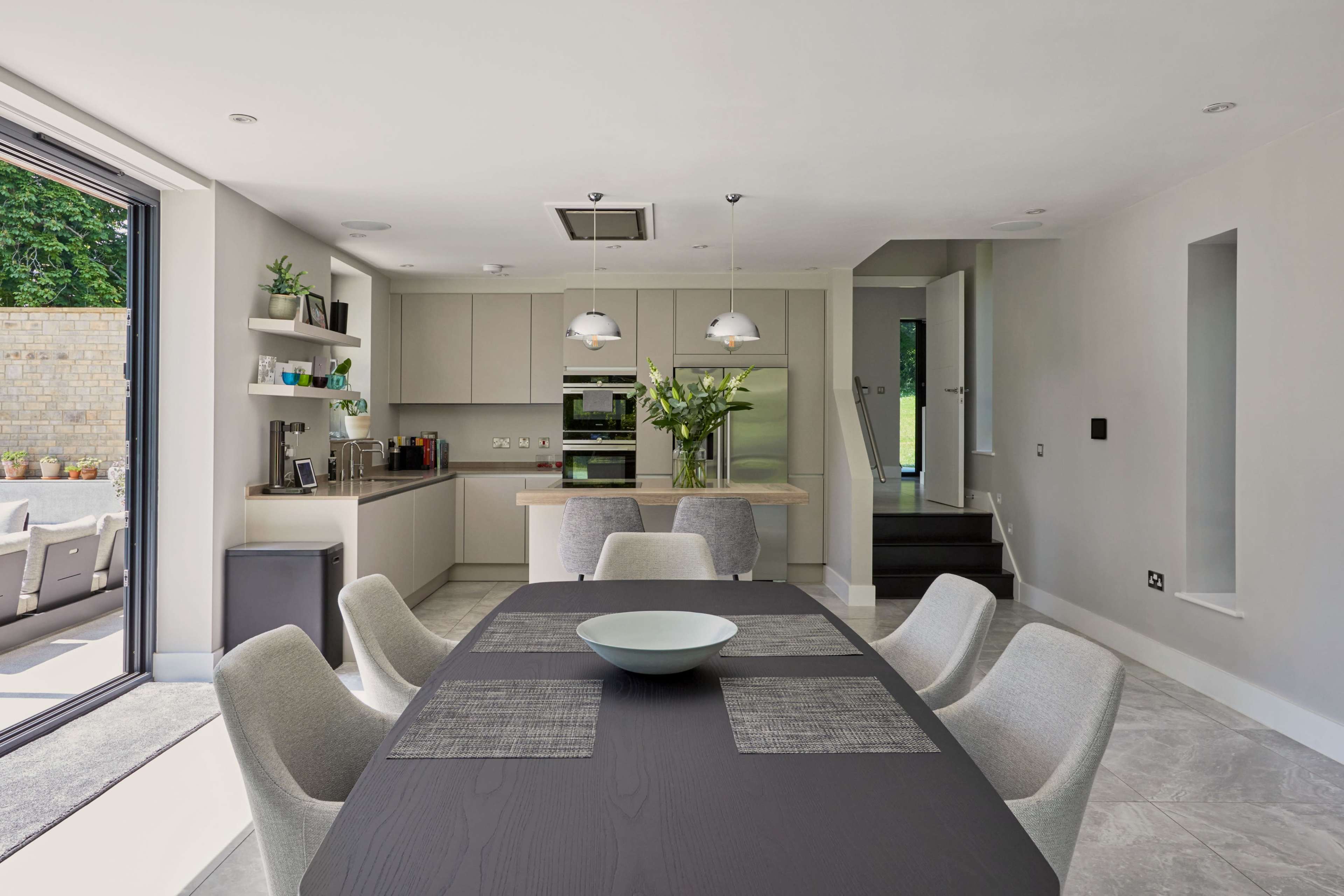 The image shows a modern kitchen and dining area featuring a large black table with gray upholstered chairs, a light-colored cabinetry setup, and a doorway leading to an outdoor space.