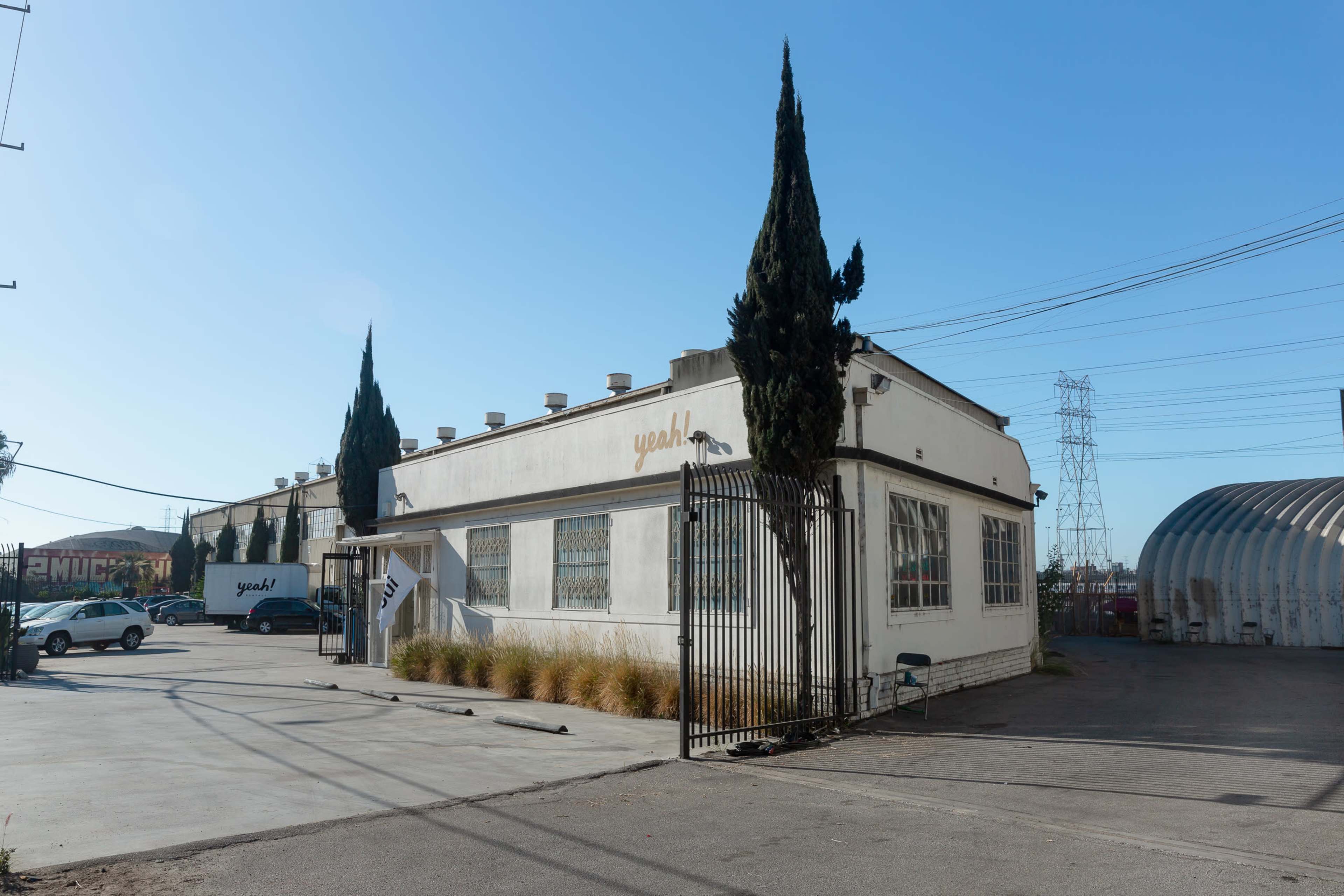 The image shows a white industrial building with a tall cypress tree in front, surrounded by a gated area and parking lot under a clear blue sky.
