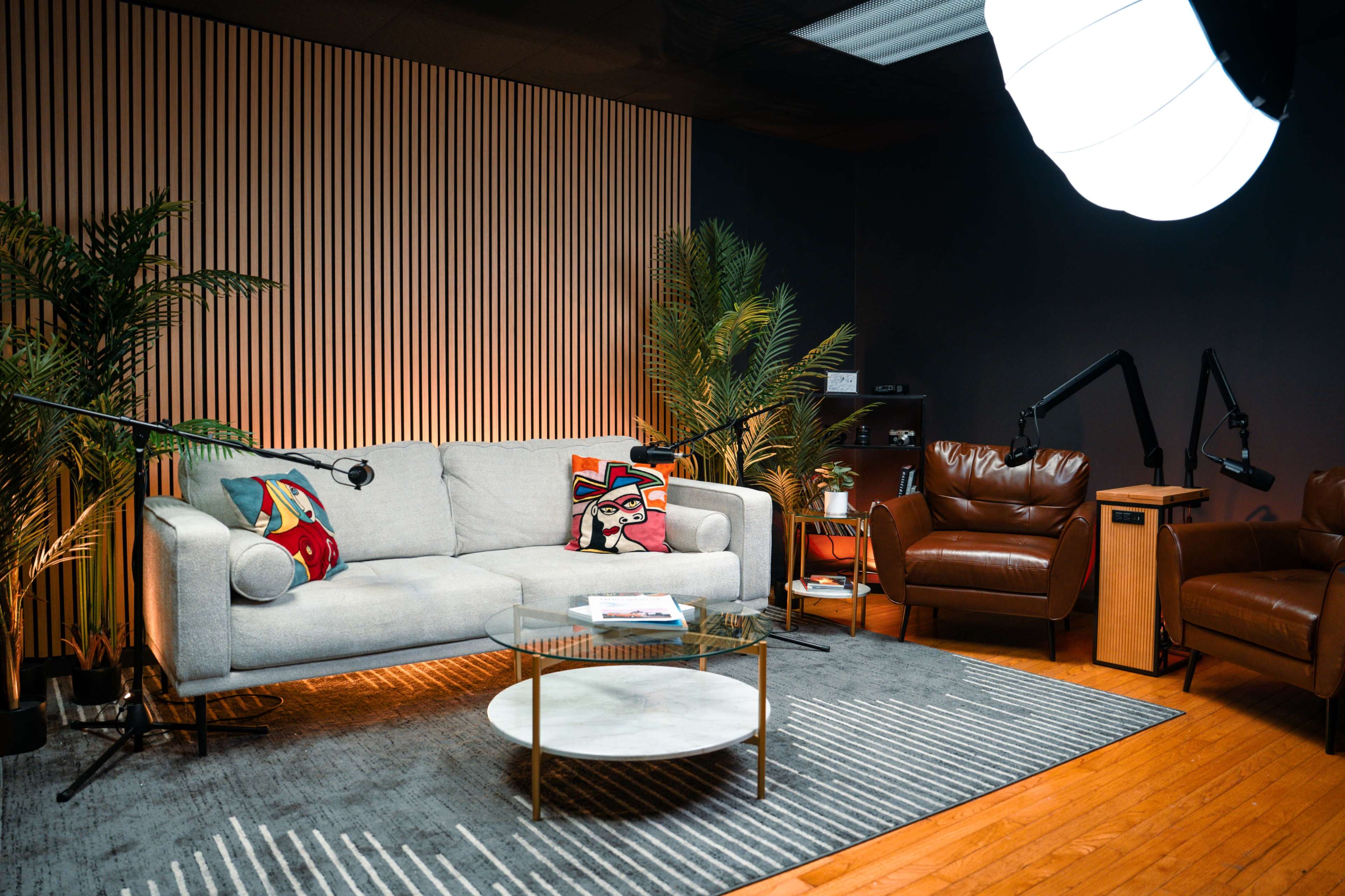 The image shows a modern living room setup with a gray sofa, two brown leather chairs, a glass coffee table, and various indoor plants against a backdrop of wooden paneling.