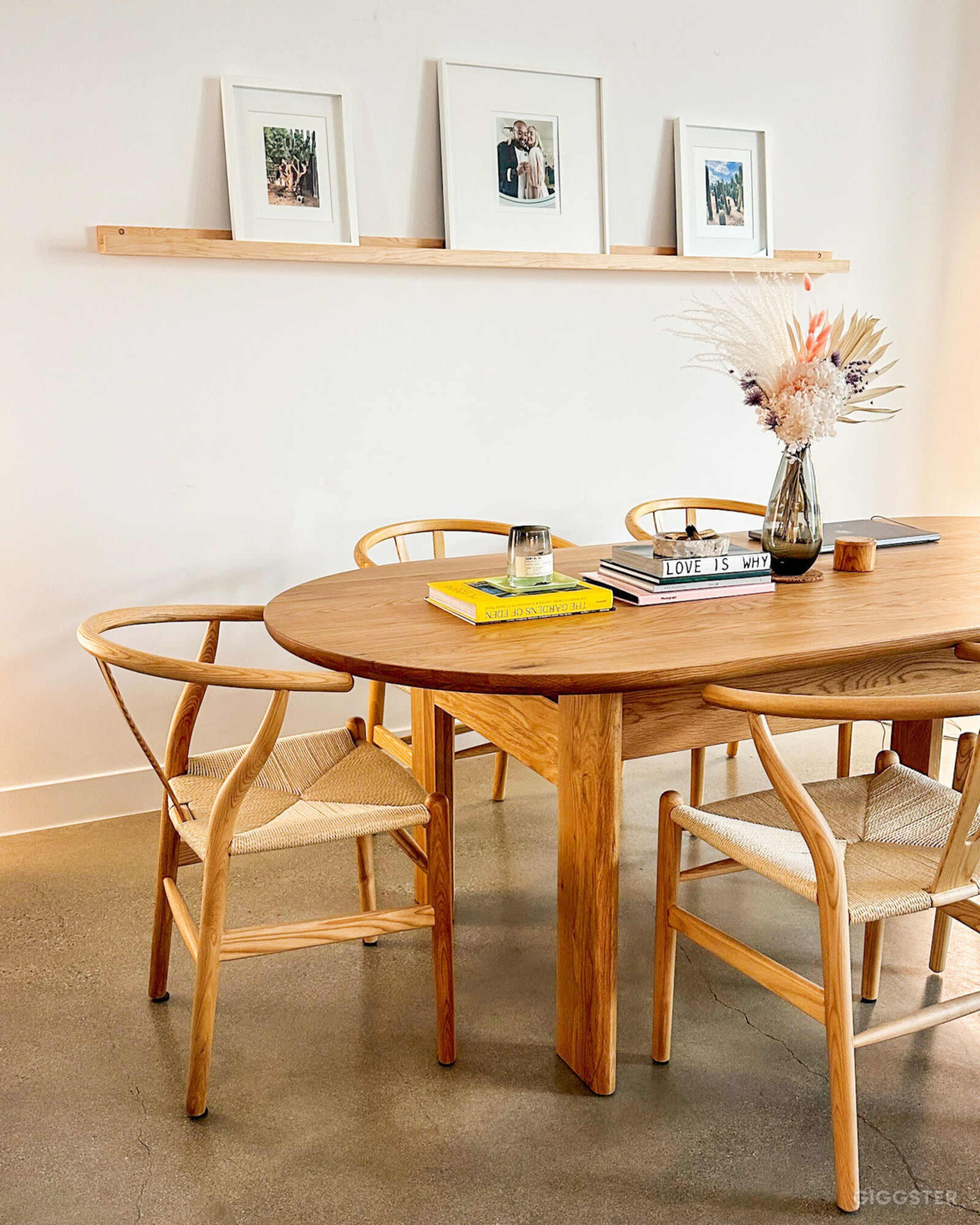 A wooden dining table with four chairs surrounded by framed photographs and a vase of dried flowers on a light-colored wall.