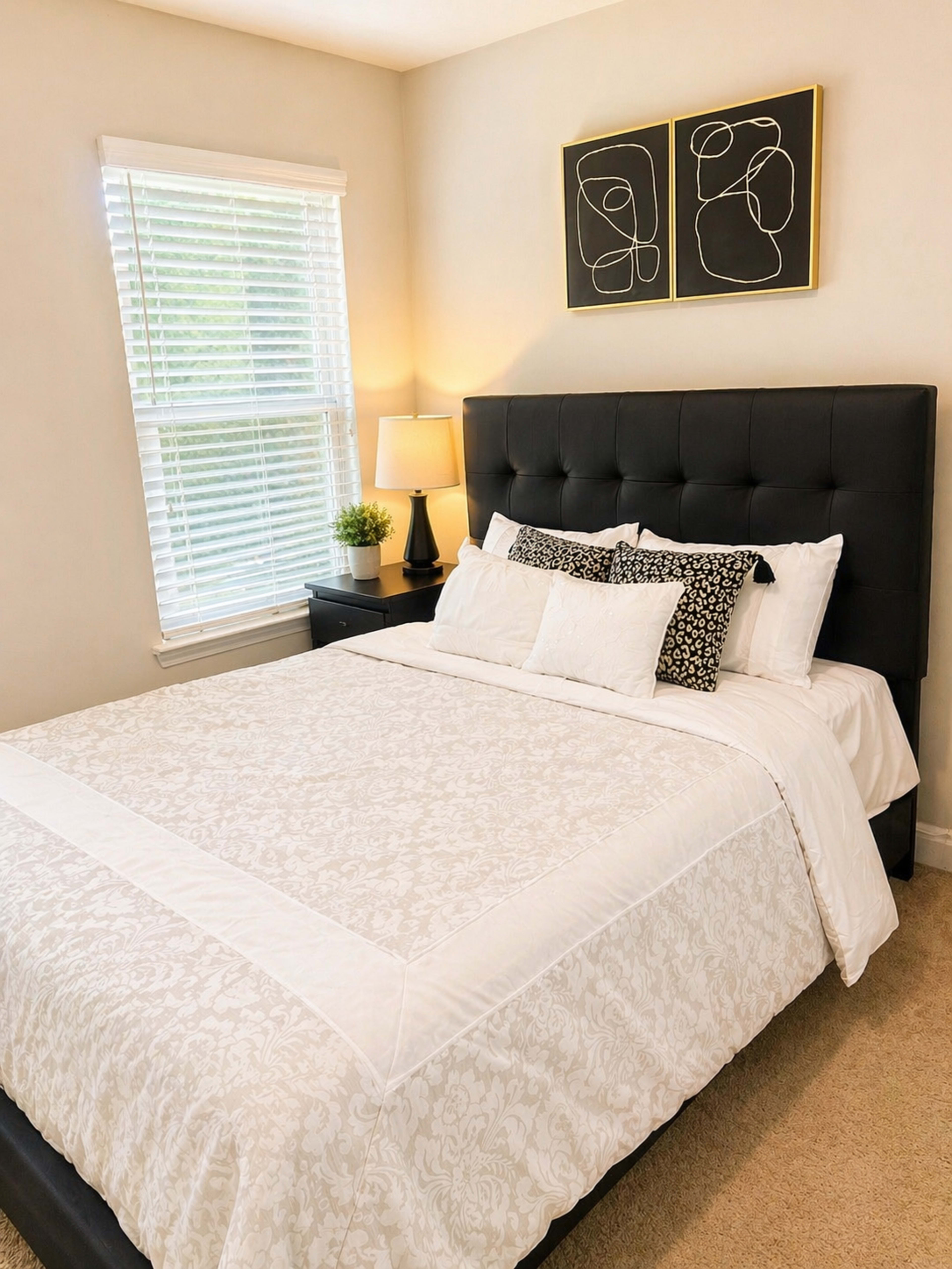 The image shows a neatly made bed with a white patterned bedspread, black headboard, and decorative pillows, positioned next to a bedside table with a lamp and a small plant.