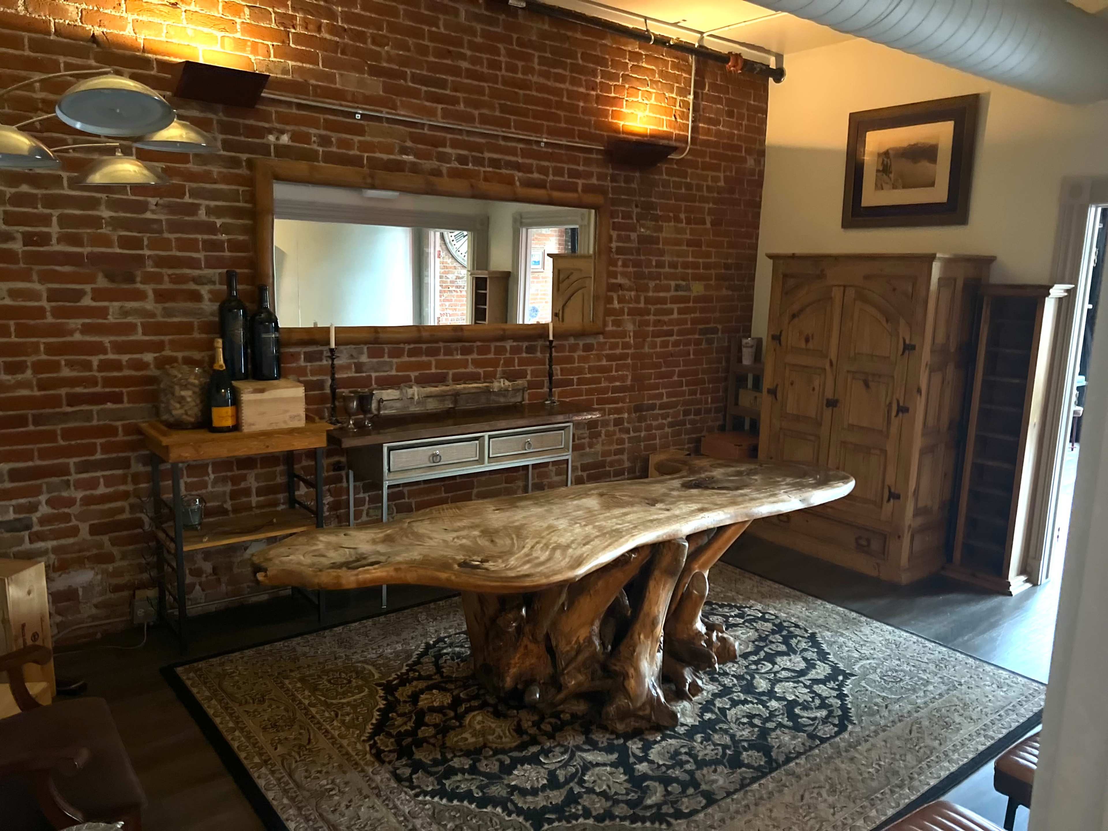 A room with exposed brick walls featuring a wooden table with a natural edge, a metal and wood side table, a rustic cabinet, and a large area rug.