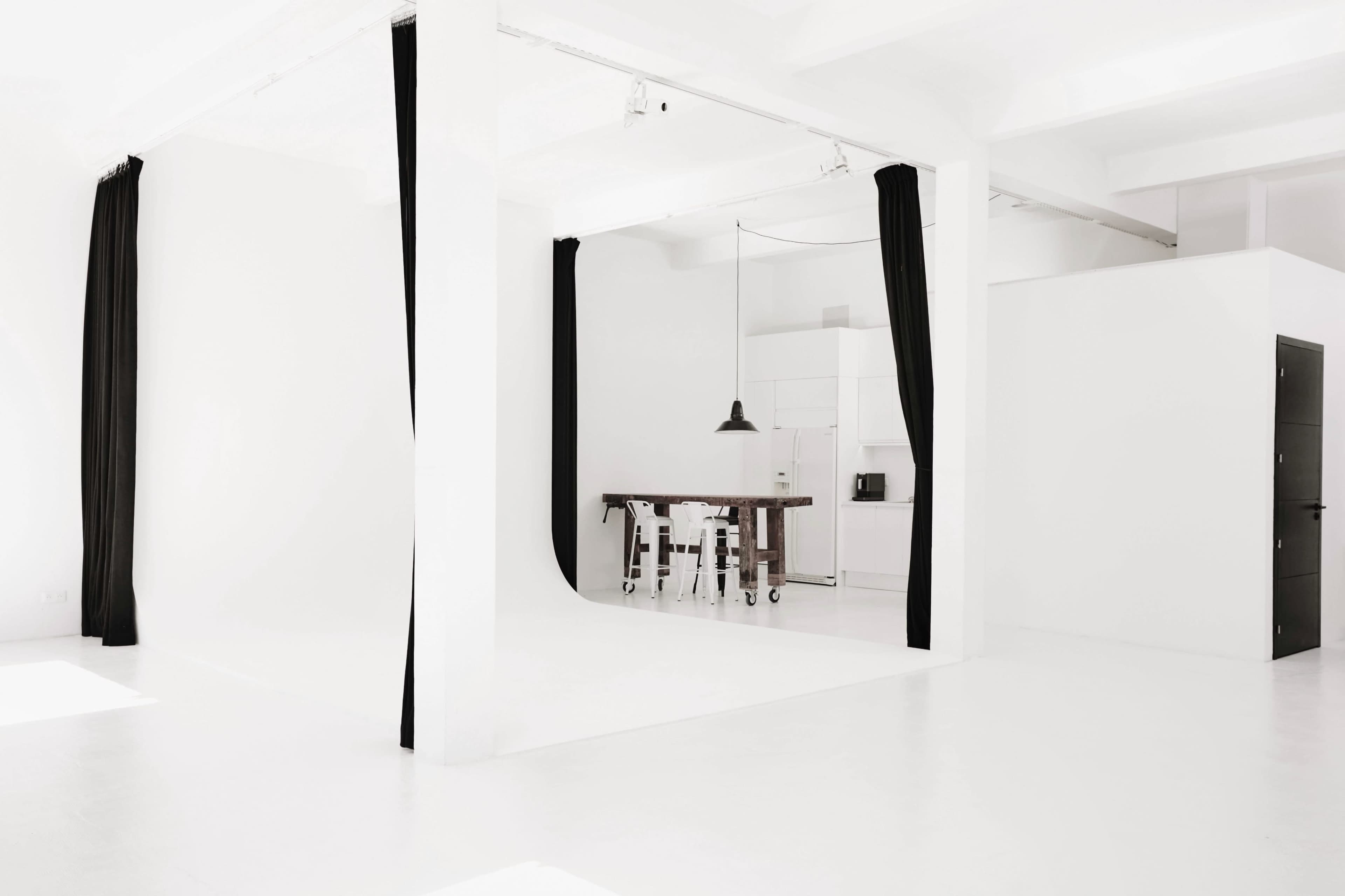 A minimalist studio space with a white interior, featuring a wooden table and black curtains.