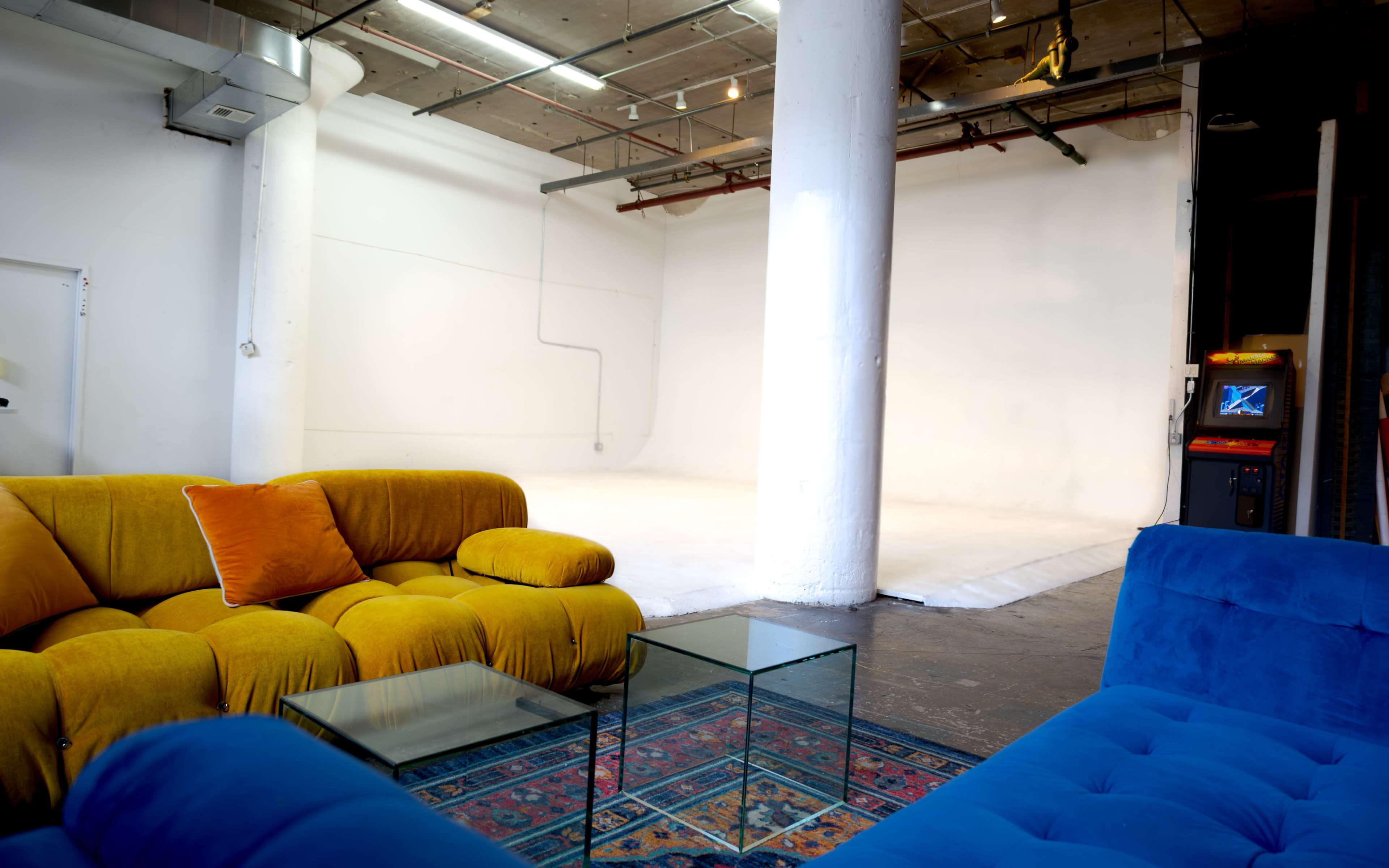 The image shows a bright studio space with a white backdrop, two colorful sofas in yellow and blue, a glass coffee table, and an arcade machine in the background.