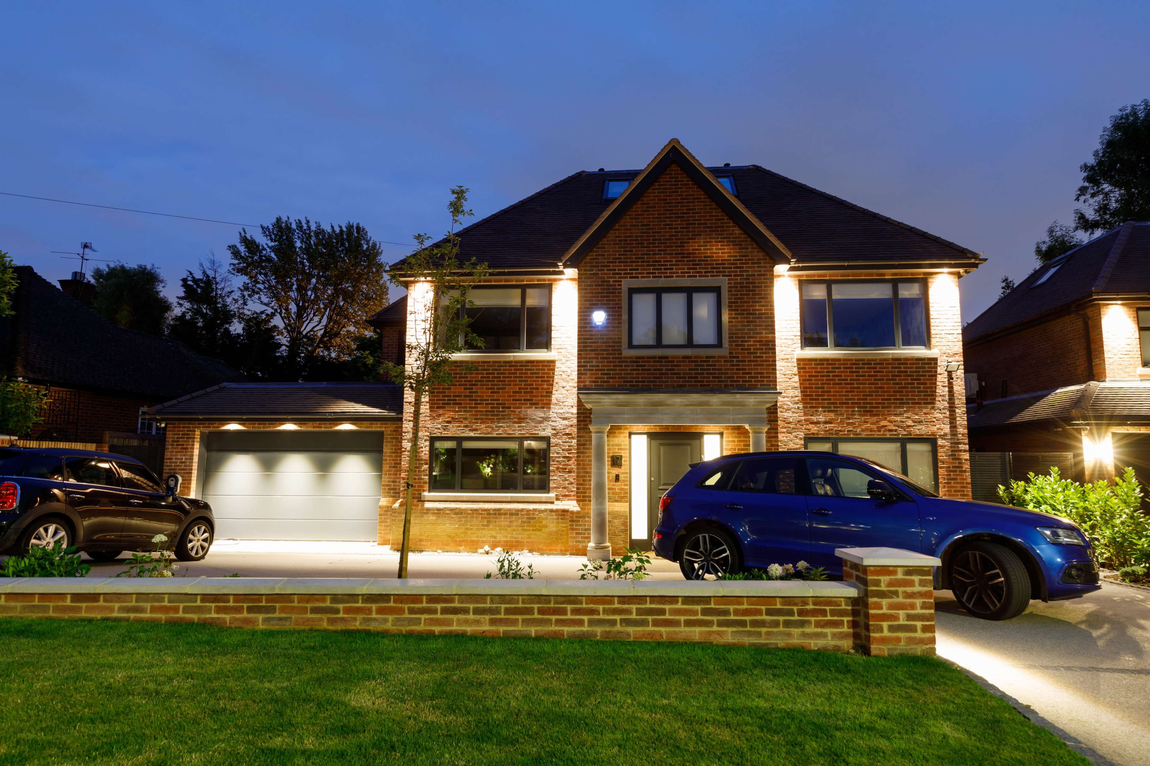 A modern two-story brick house is illuminated at night, featuring a front yard and a driveway with parked cars.