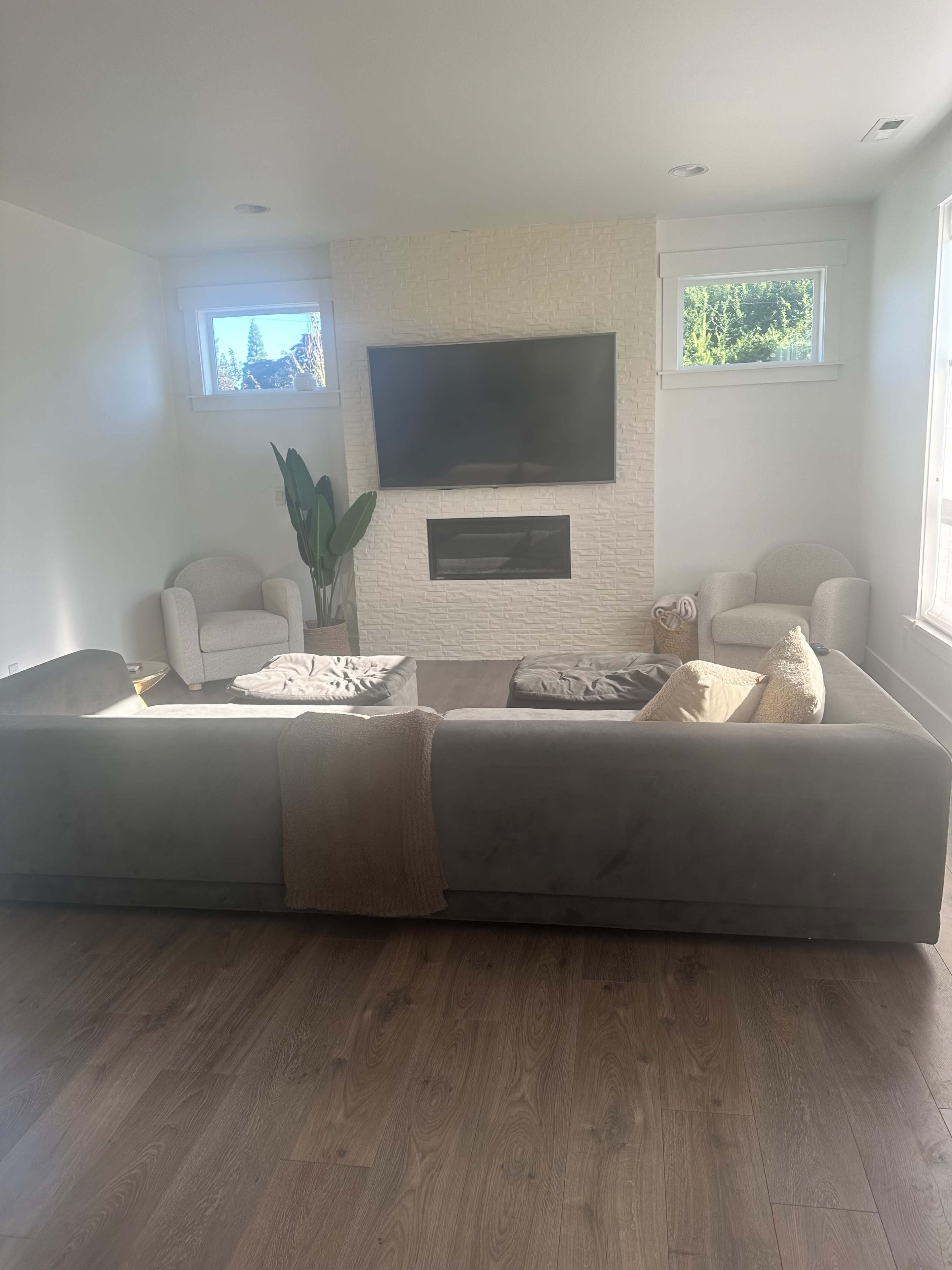 A modern living room features a large sectional sofa facing a wall-mounted television above a fireplace, with two armchairs and a potted plant in the corner.