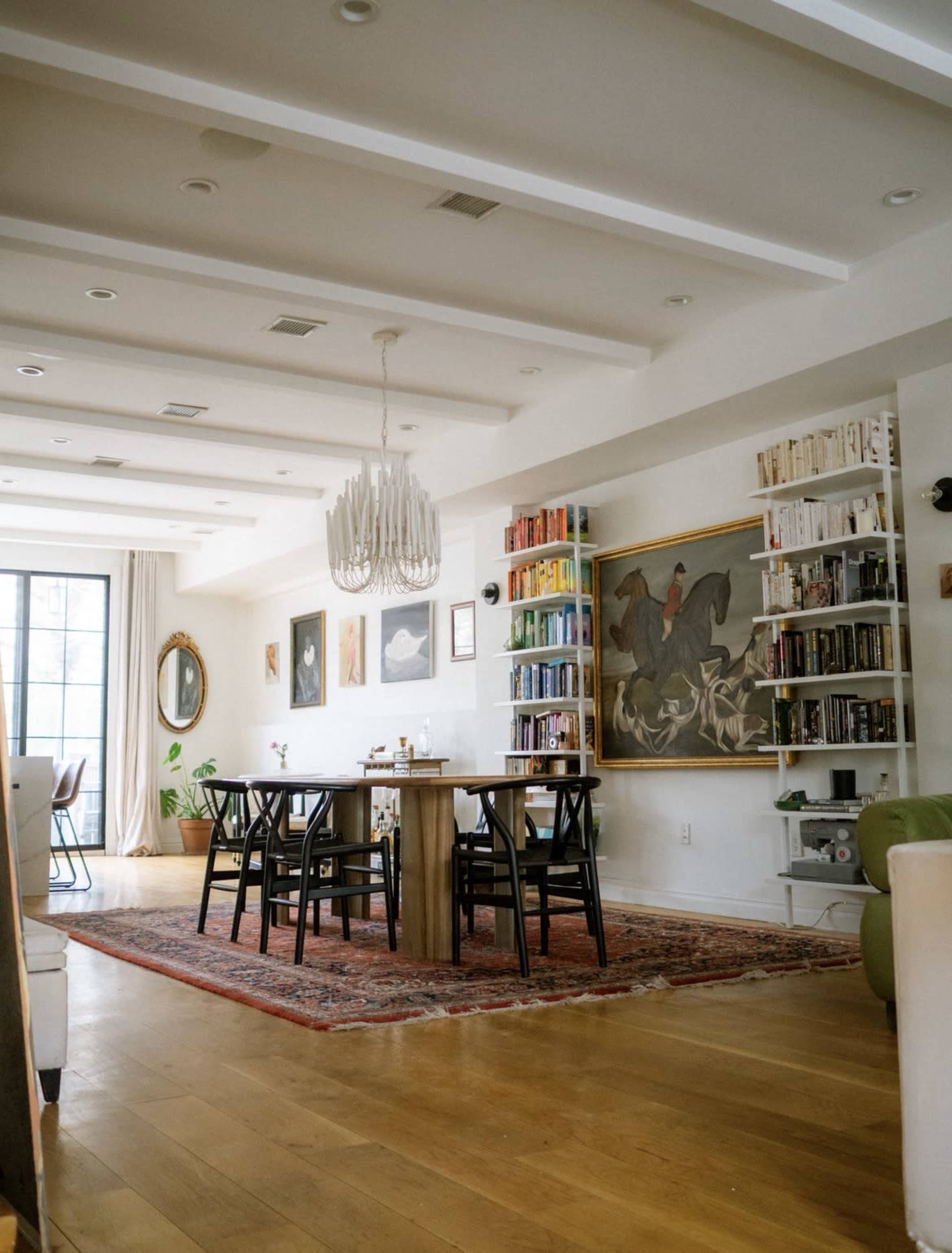The image shows a spacious, well-lit living area with a dining table, bookshelves filled with colorful books, and framed artwork on the walls.