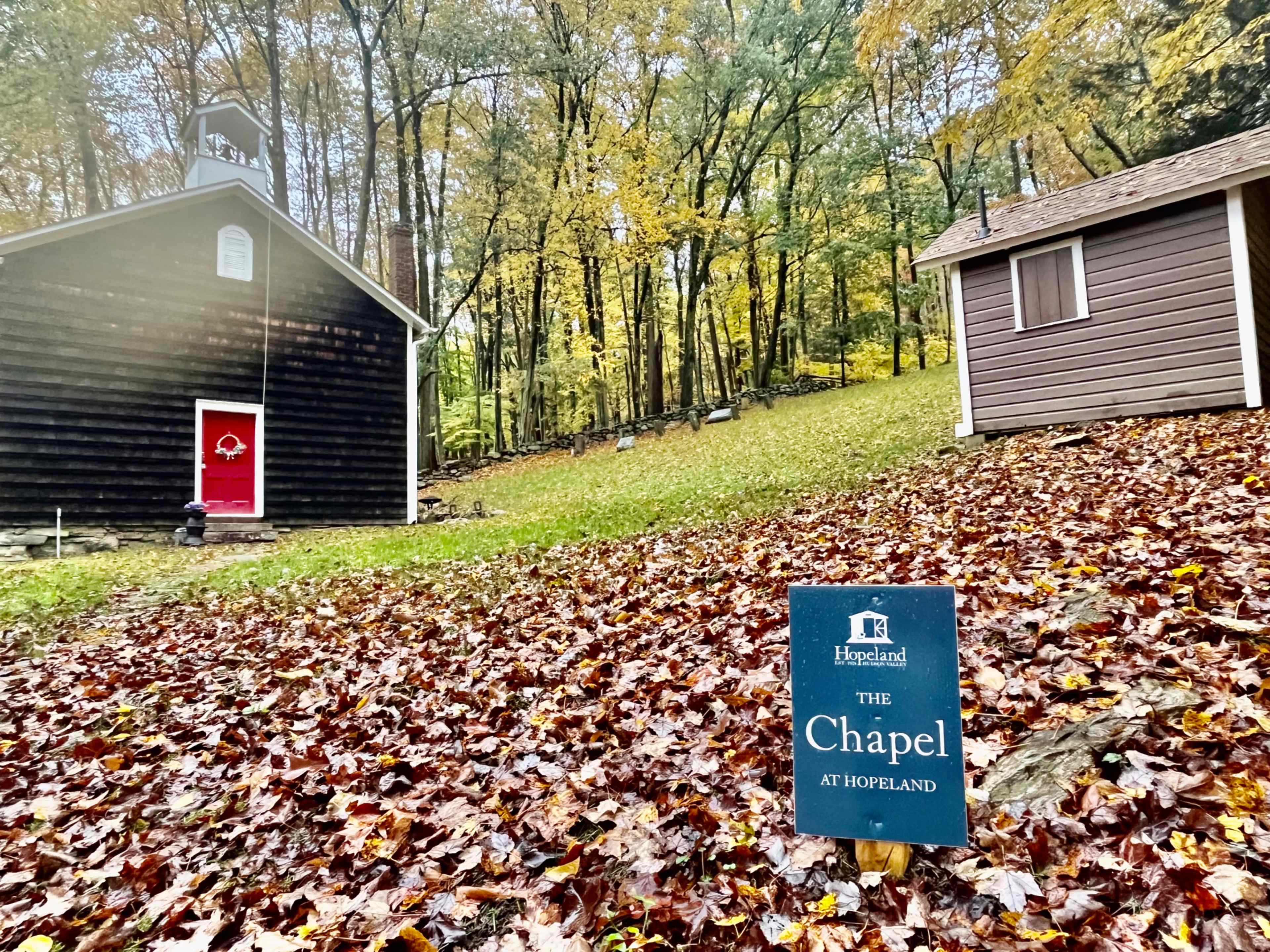 The image shows a small chapel building with a red door alongside a modern cabin, both surrounded by autumn foliage on the ground.