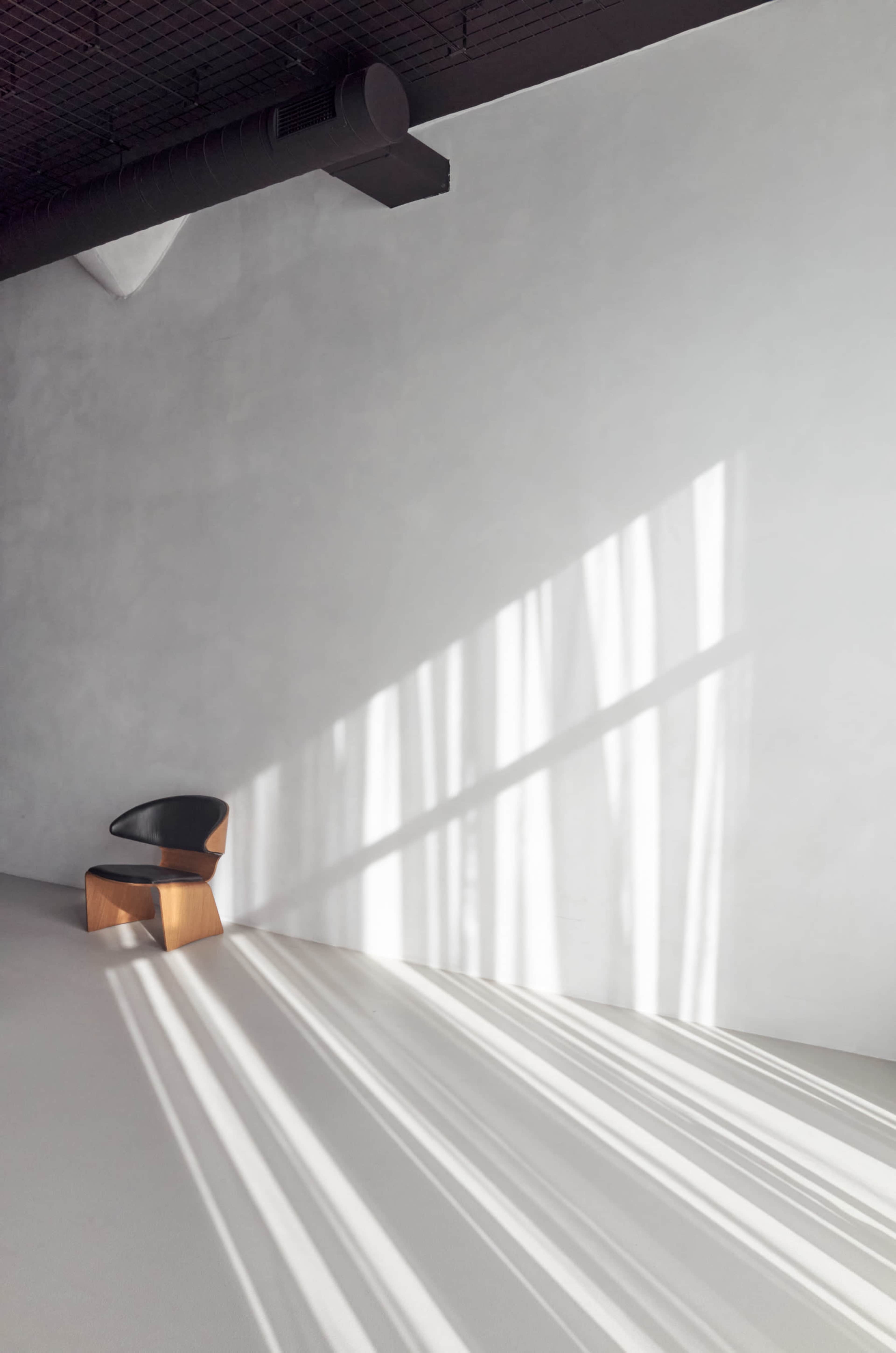A minimalist interior features a black chair with a wooden base positioned against a pale wall, while long shadows from nearby windows stretch across the floor.