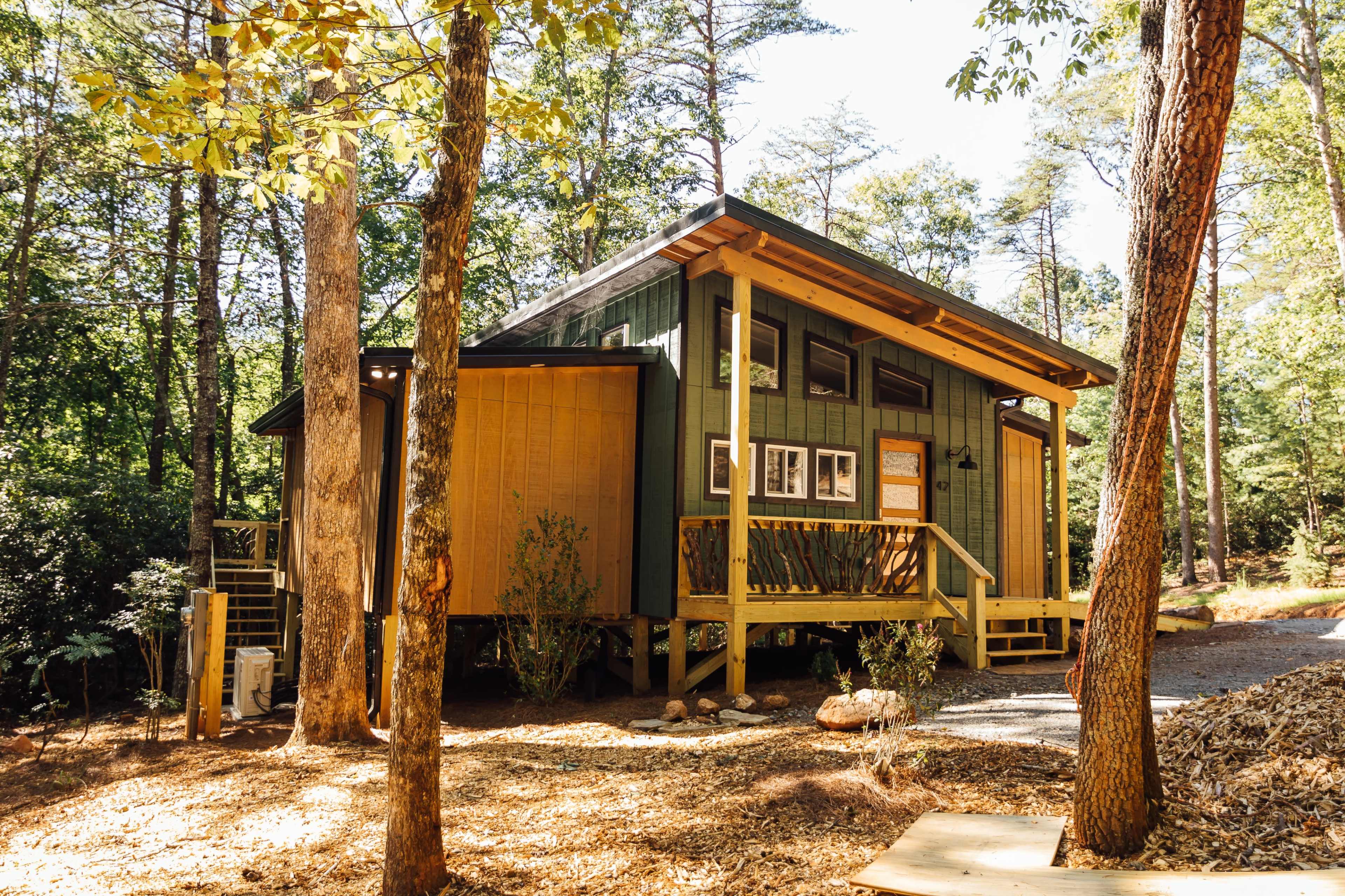 A green and yellow cabin is surrounded by trees and features a wooden deck and porch.