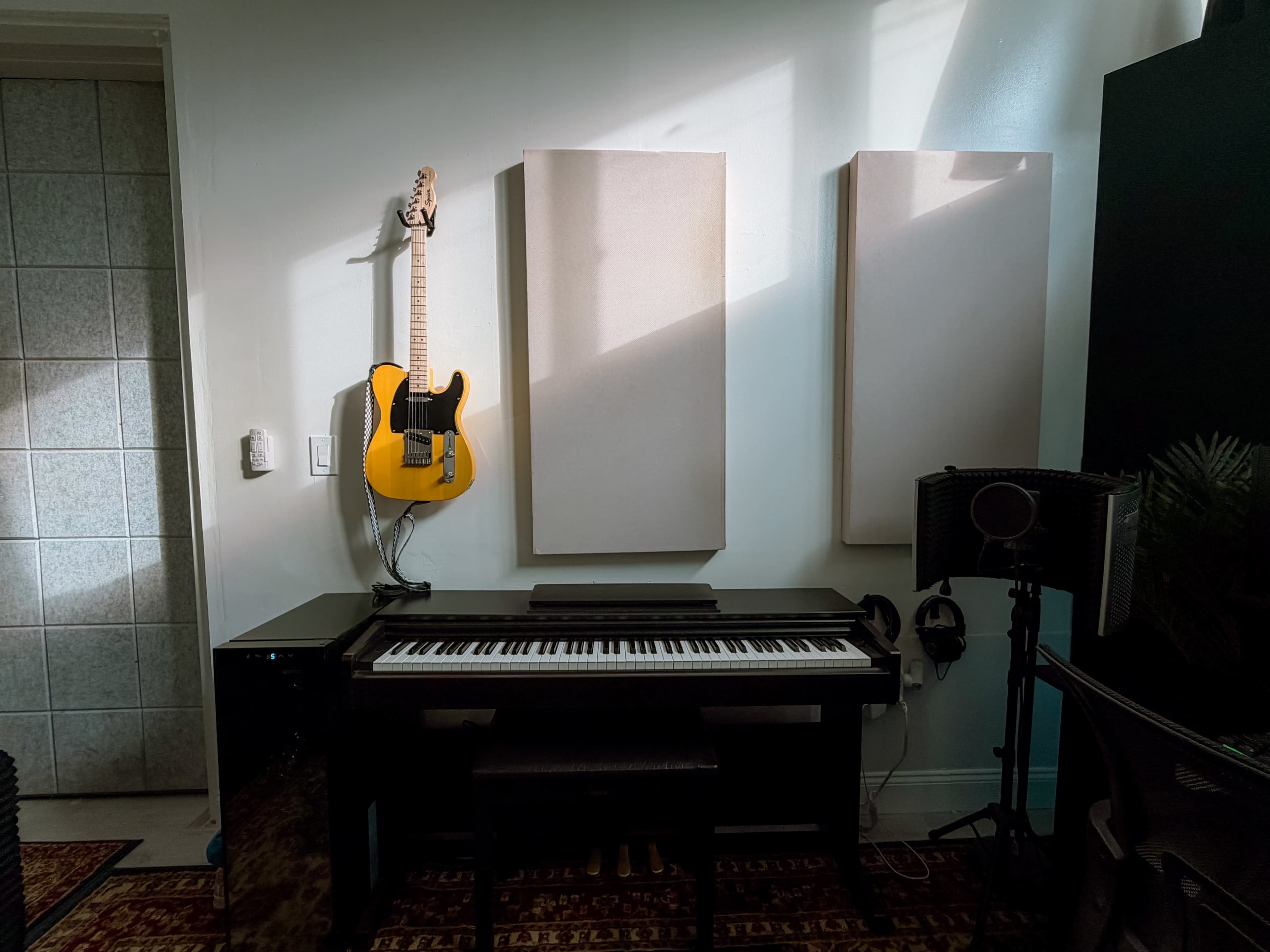 The image shows a corner of a music studio featuring a digital piano, a bright yellow guitar mounted on the wall, and acoustic panels.