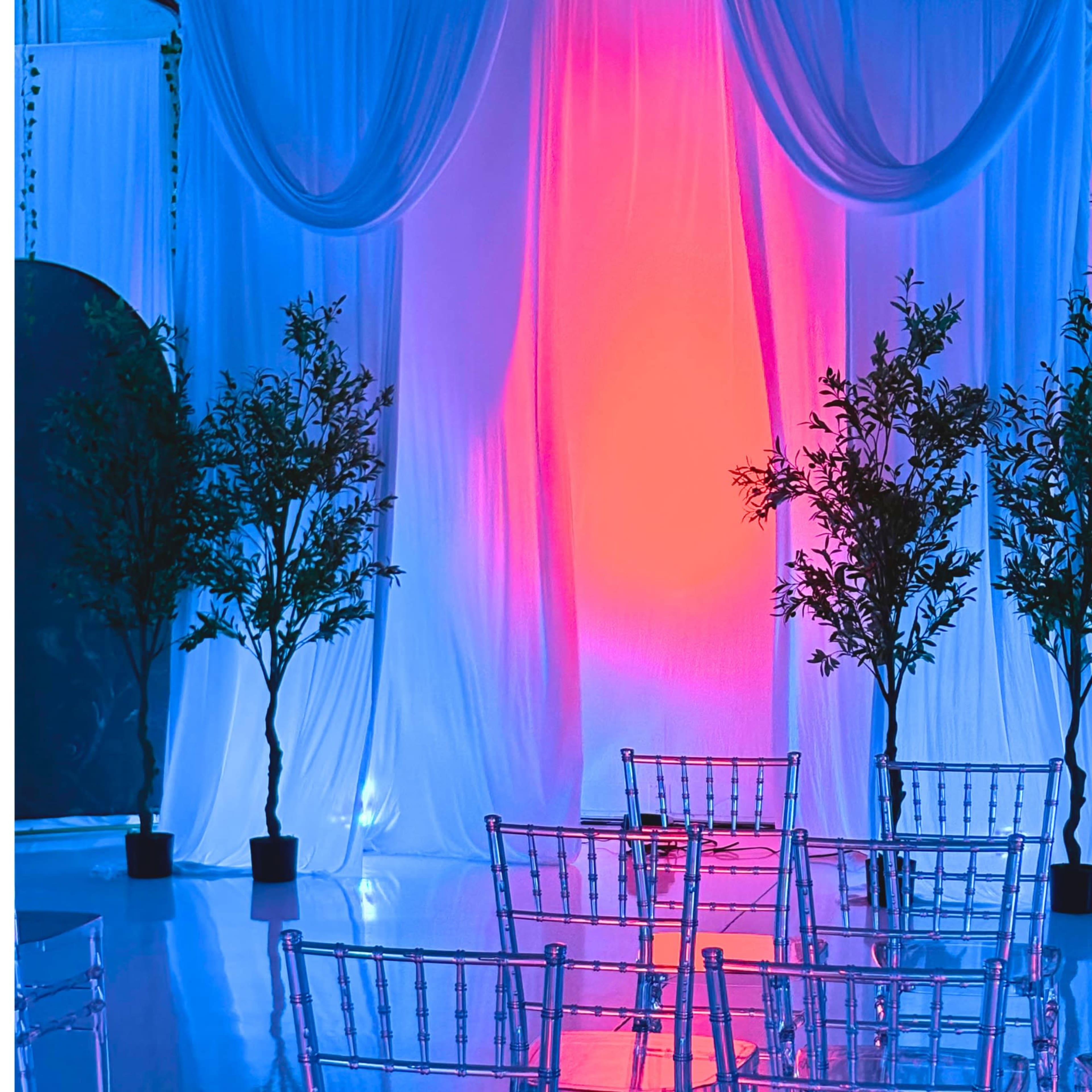 The scene features a decorated indoor space with blue and pink lighting, draped fabric as a backdrop, artificial trees, and clear plastic chairs arranged in rows.