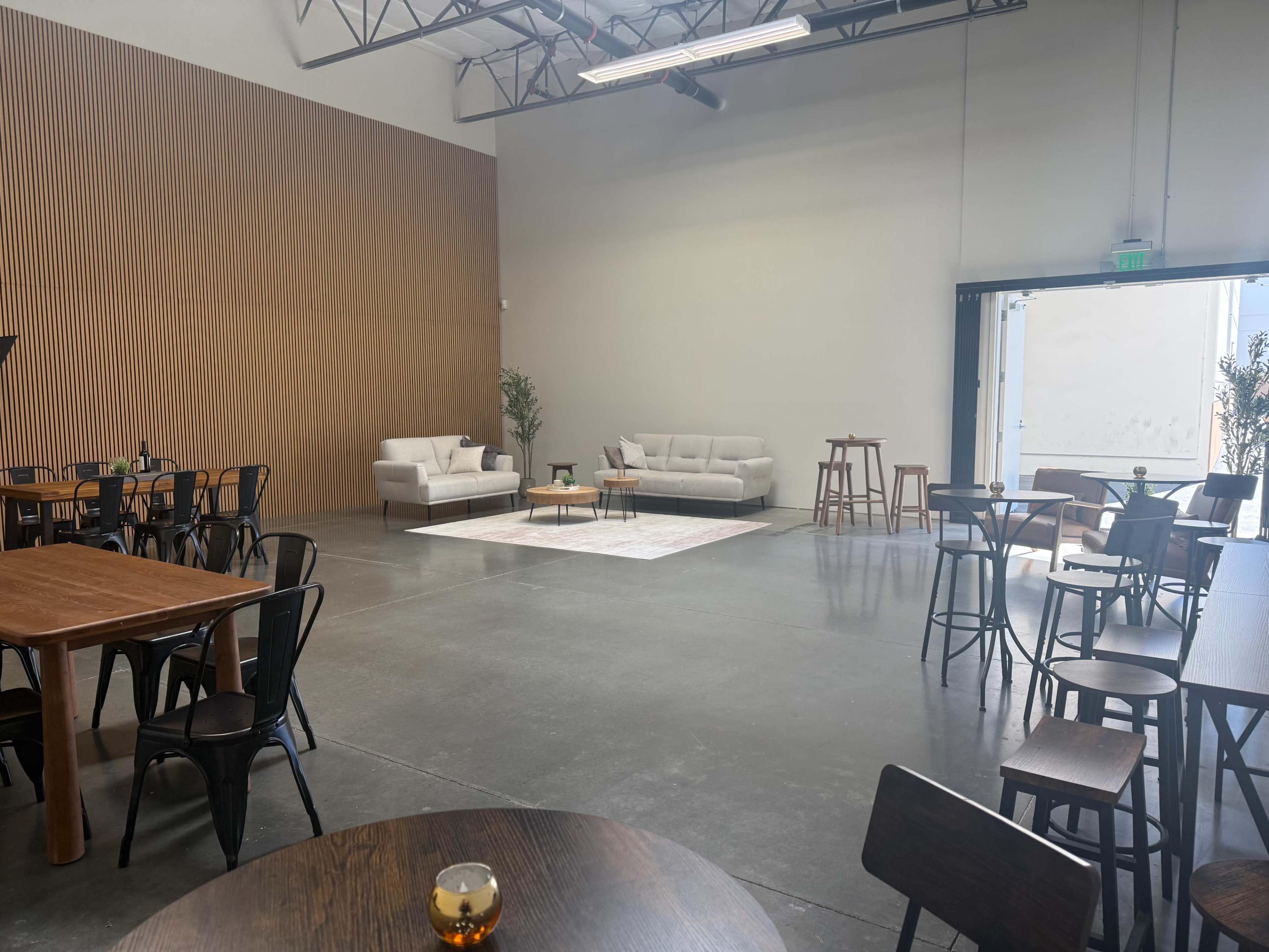 The image shows a spacious indoor area with wooden tables, black chairs, a coffee table on a rug, and seating arrangements including two sofas near an open garage door.