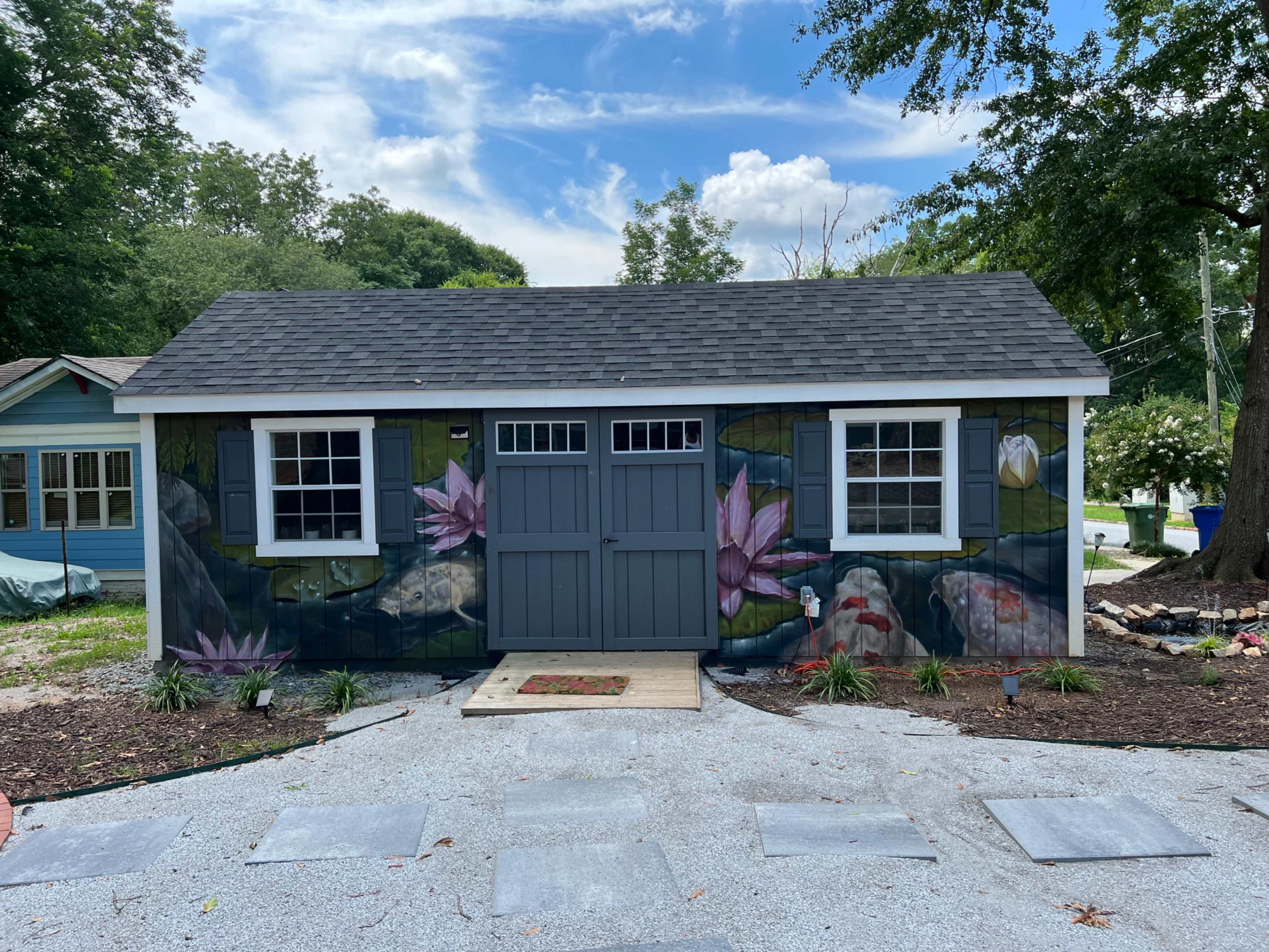 A garden shed with a mural of water lilies and koi fish is surrounded by a gravel pathway and landscaped greenery.