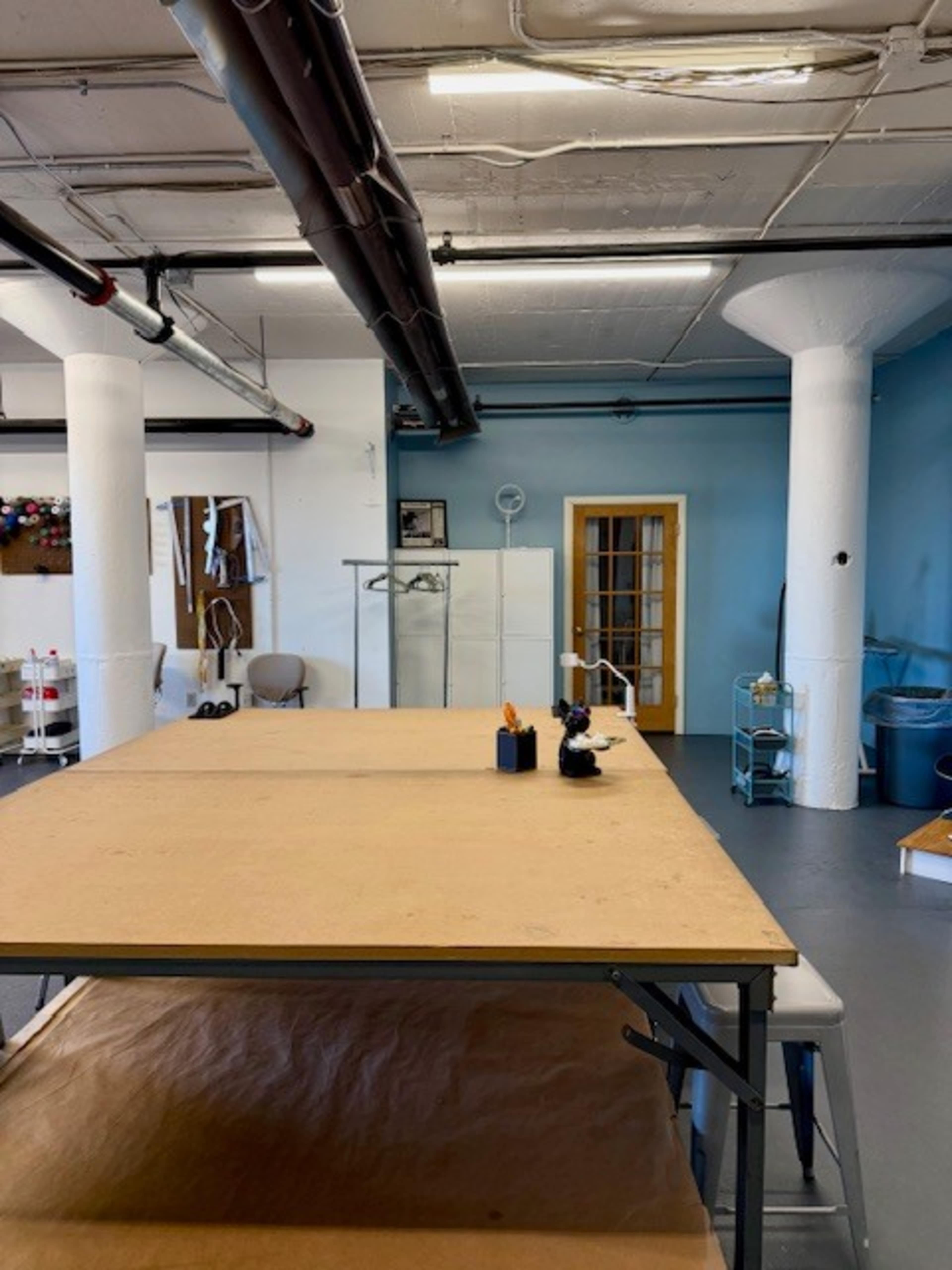 The image shows a spacious workshop with a large wooden table in the foreground, white pillars, and light blue walls, along with various tools and equipment scattered around the room.