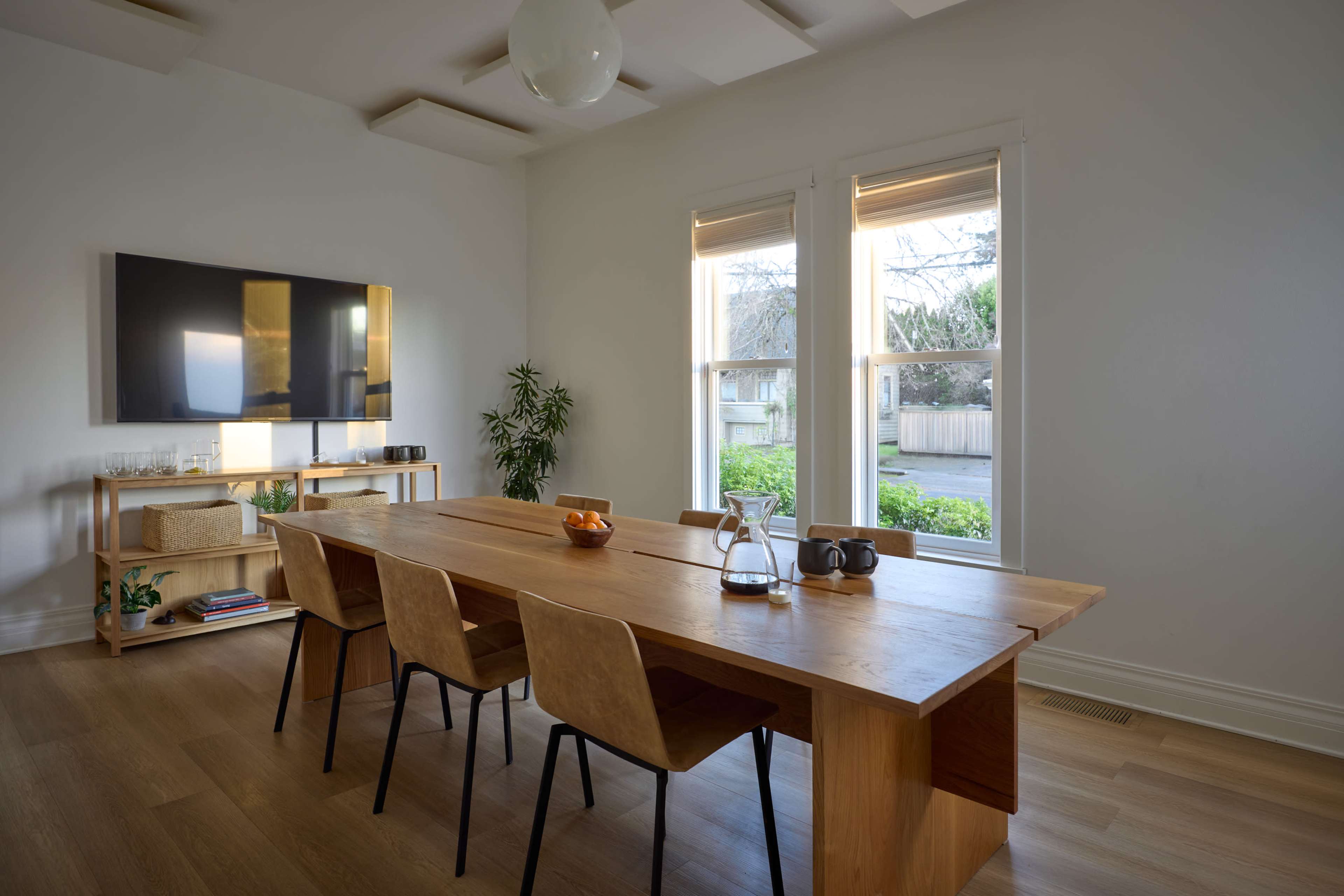 The image shows a modern dining room with a long wooden table and four chairs, a large flat-screen TV mounted on the wall, and two windows overlooking a green outdoor space.