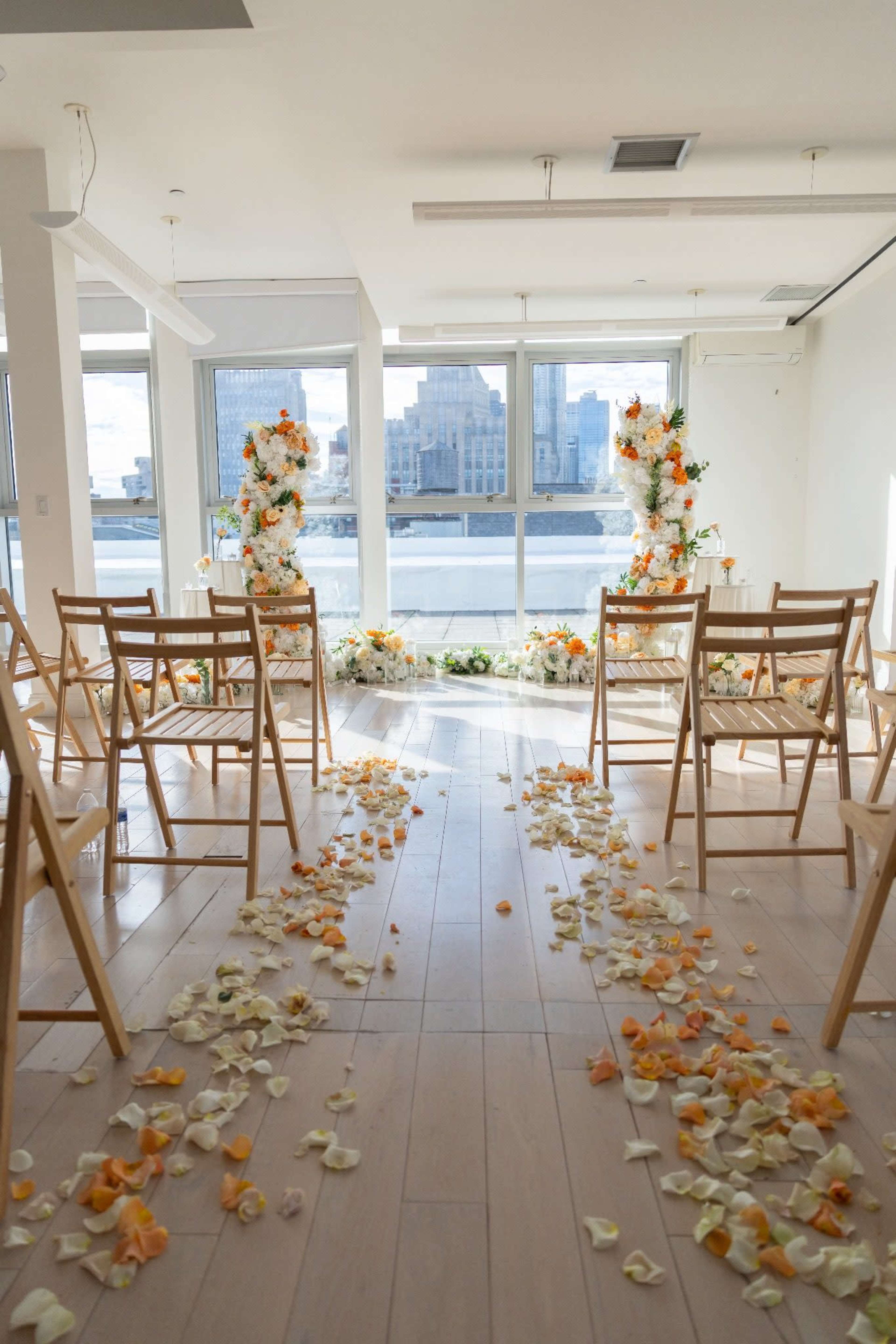 The image shows a decorated space with wooden chairs arranged in rows, flower arrangements at the front, and a path of flower petals leading to a large window with a view of buildings outside.