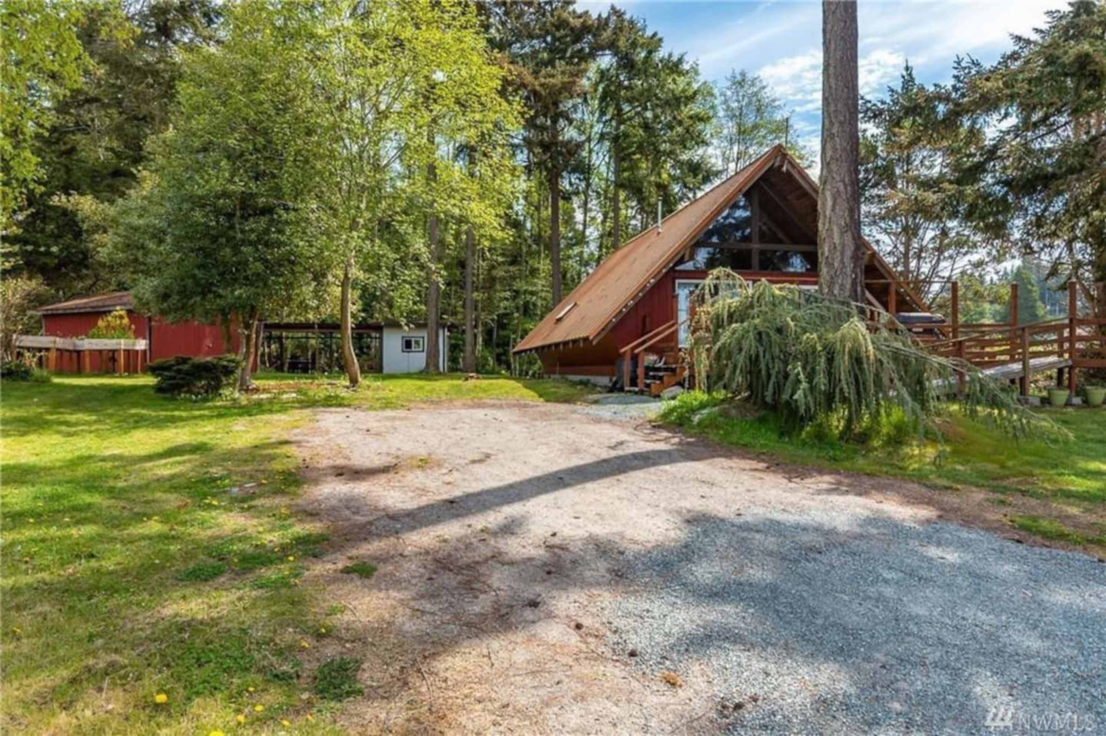 A gravel driveway leads to a tall, A-frame house surrounded by trees and a red barn in a rural setting.