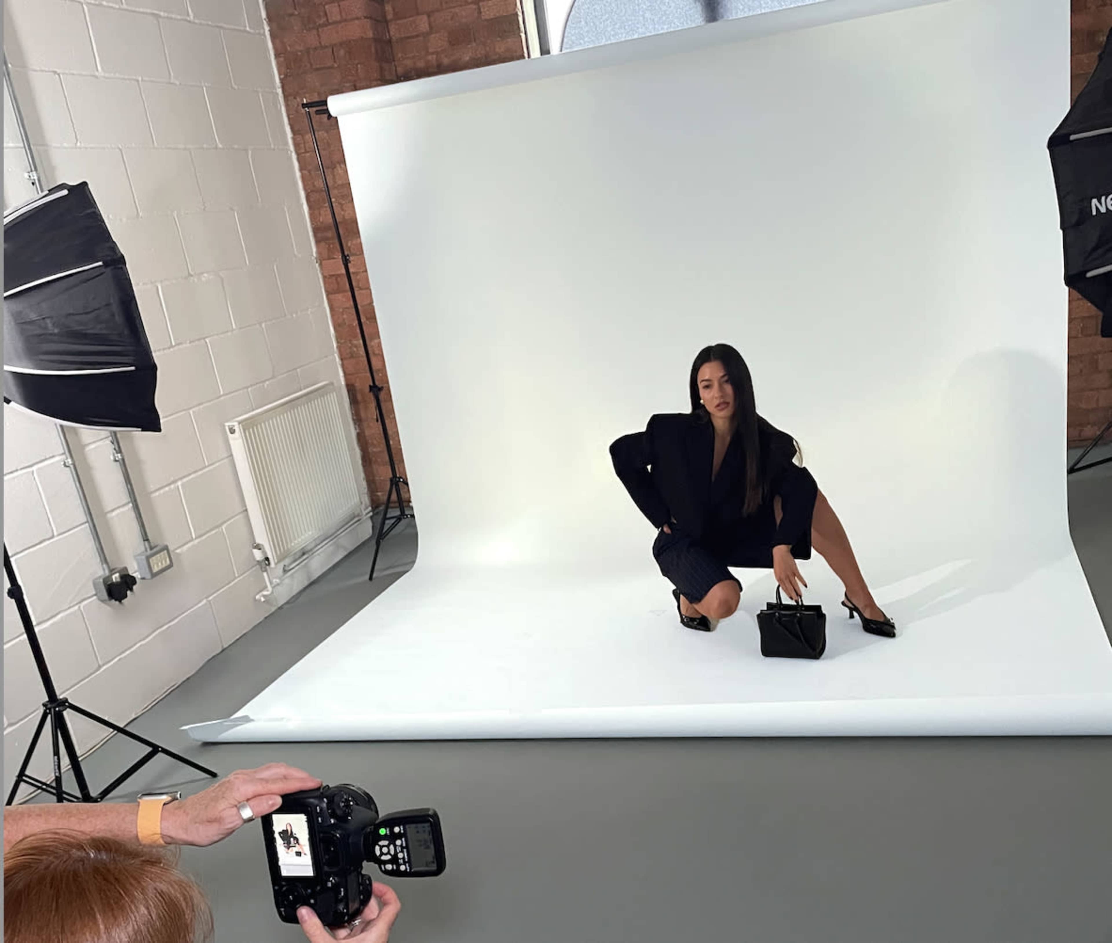 A model poses on a white backdrop during a photoshoot while a photographer captures the scene.