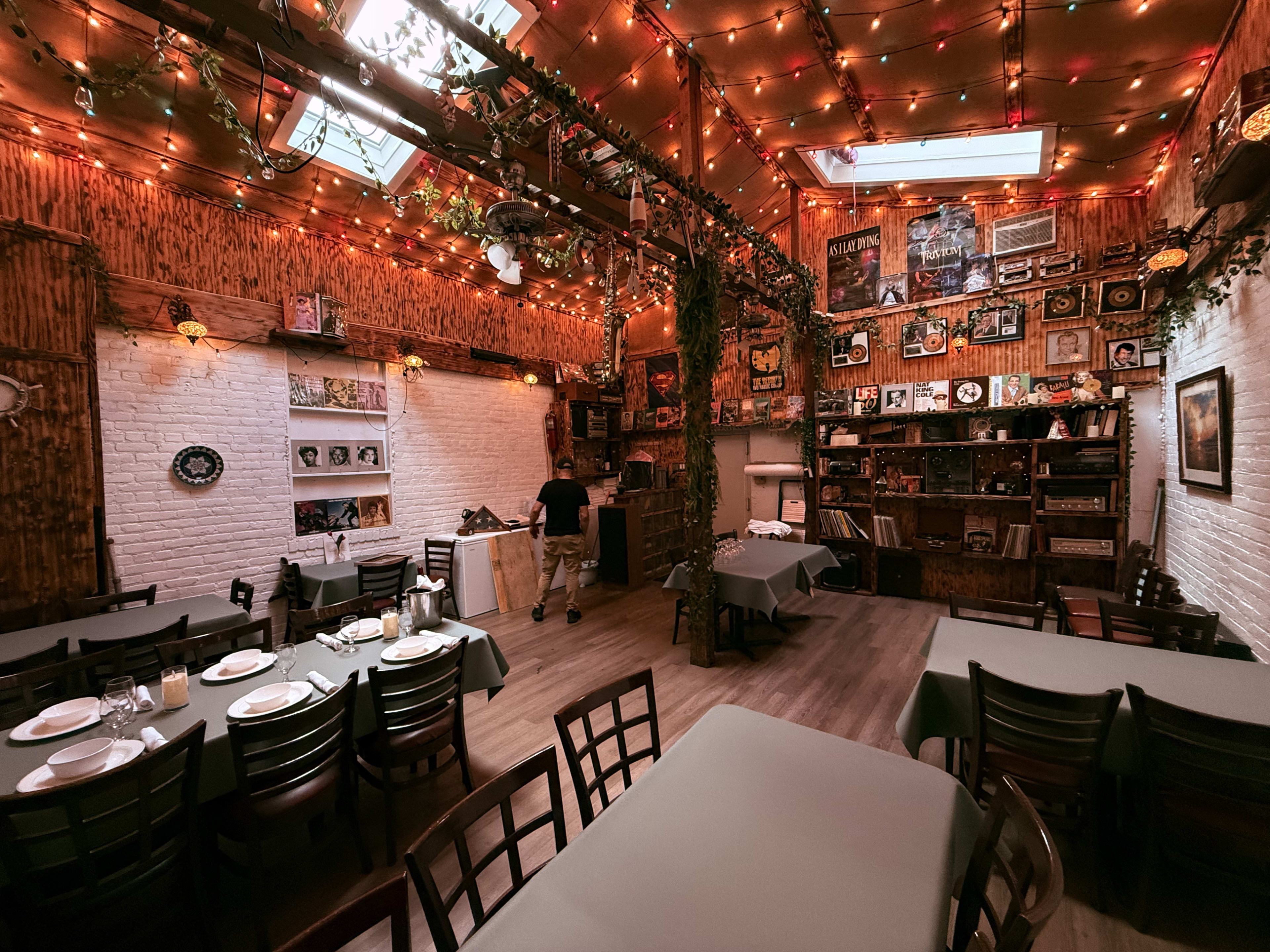 The image shows a warmly decorated restaurant interior with tables covered in green cloths, a wooden shelving unit filled with various items, and soft lighting from string lights.