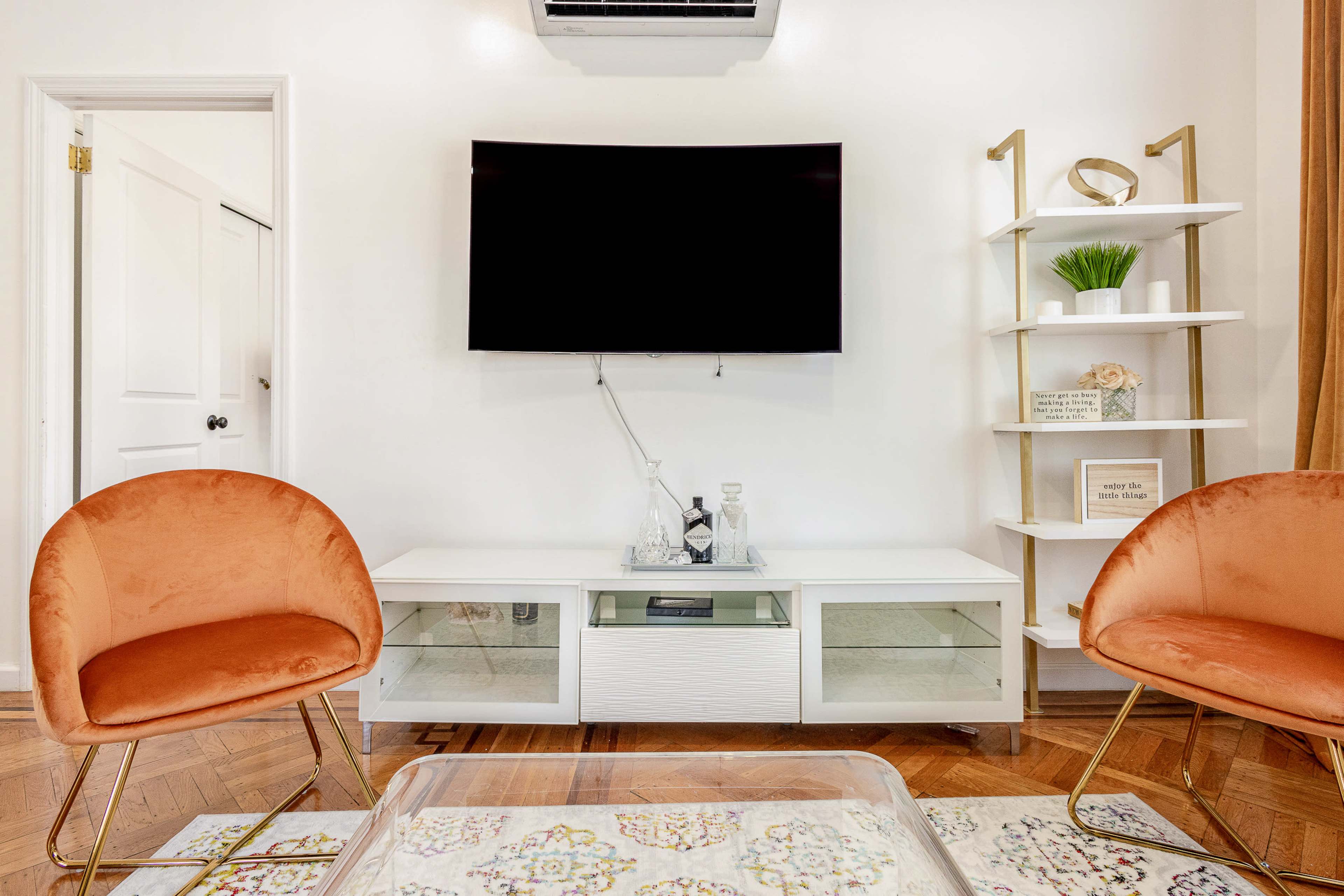 A living room with two orange chairs facing a white TV cabinet that holds a flat-screen television, accompanied by a decorative shelf and a patterned area rug.