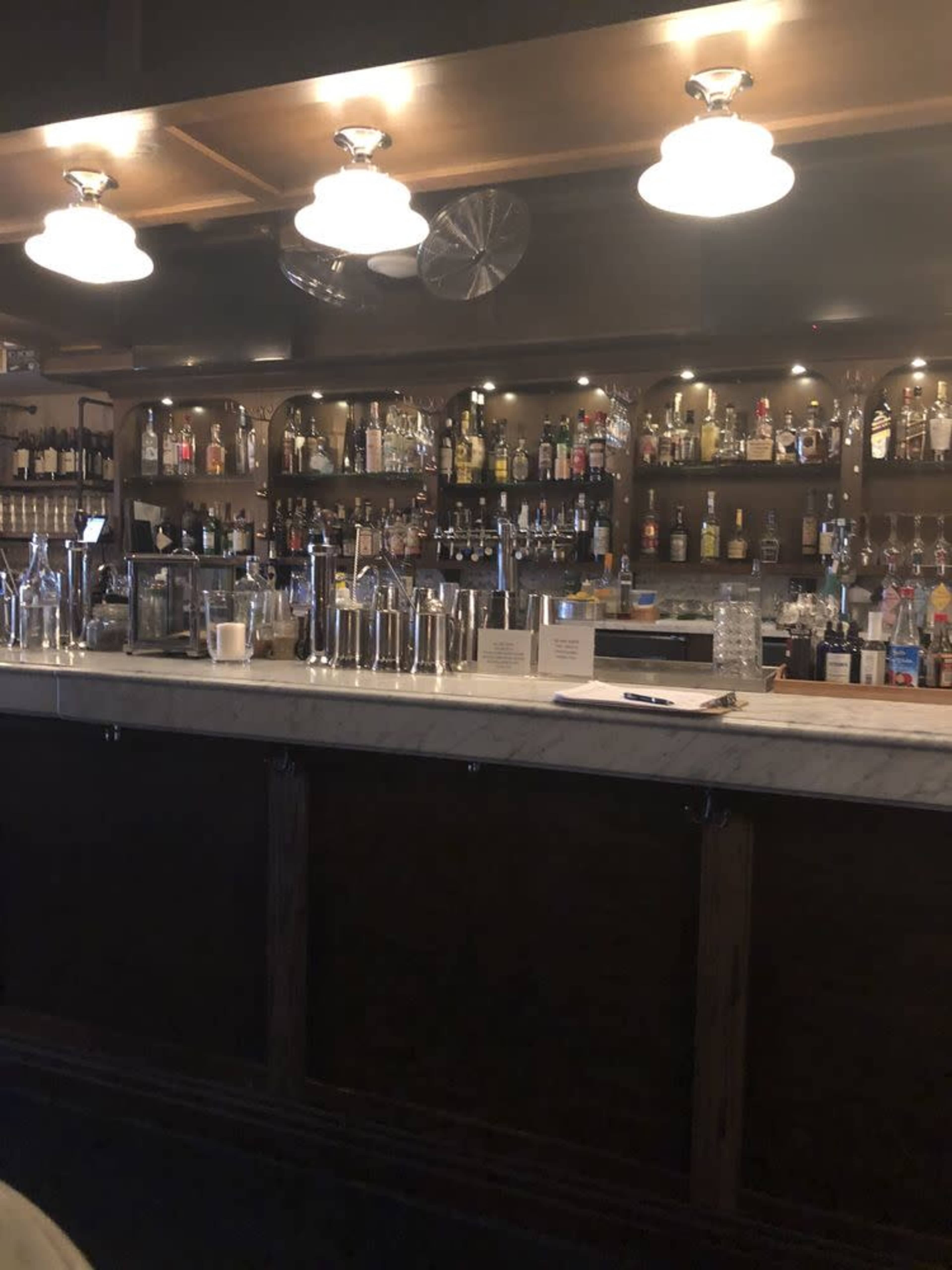 A bar counter with an array of liquor bottles and various bar tools organized neatly on a marble surface behind a wooden bar.