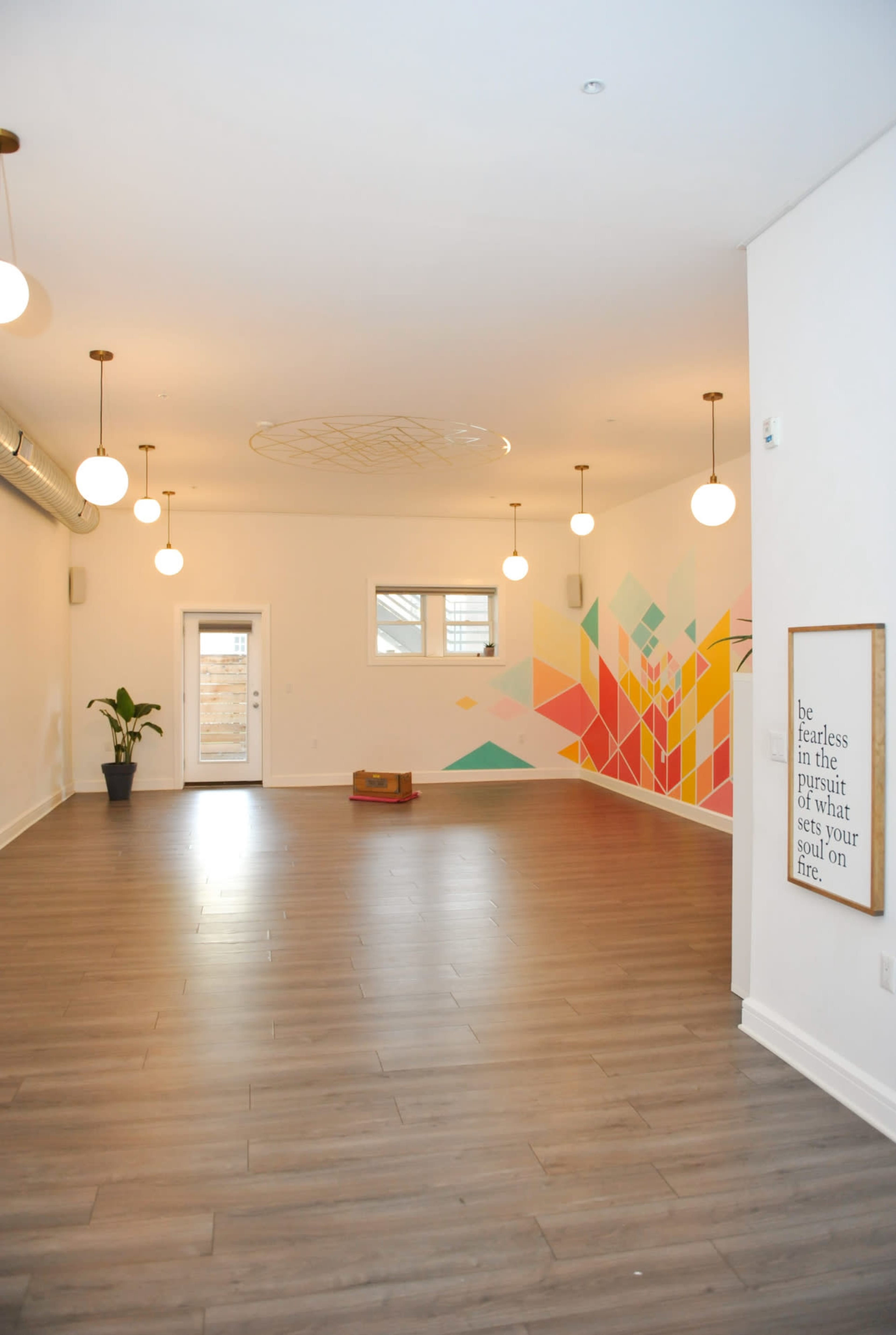 The image shows a spacious room with wooden flooring, a colorful geometric mural on the wall, several hanging lights, and a door leading to an outdoor area.