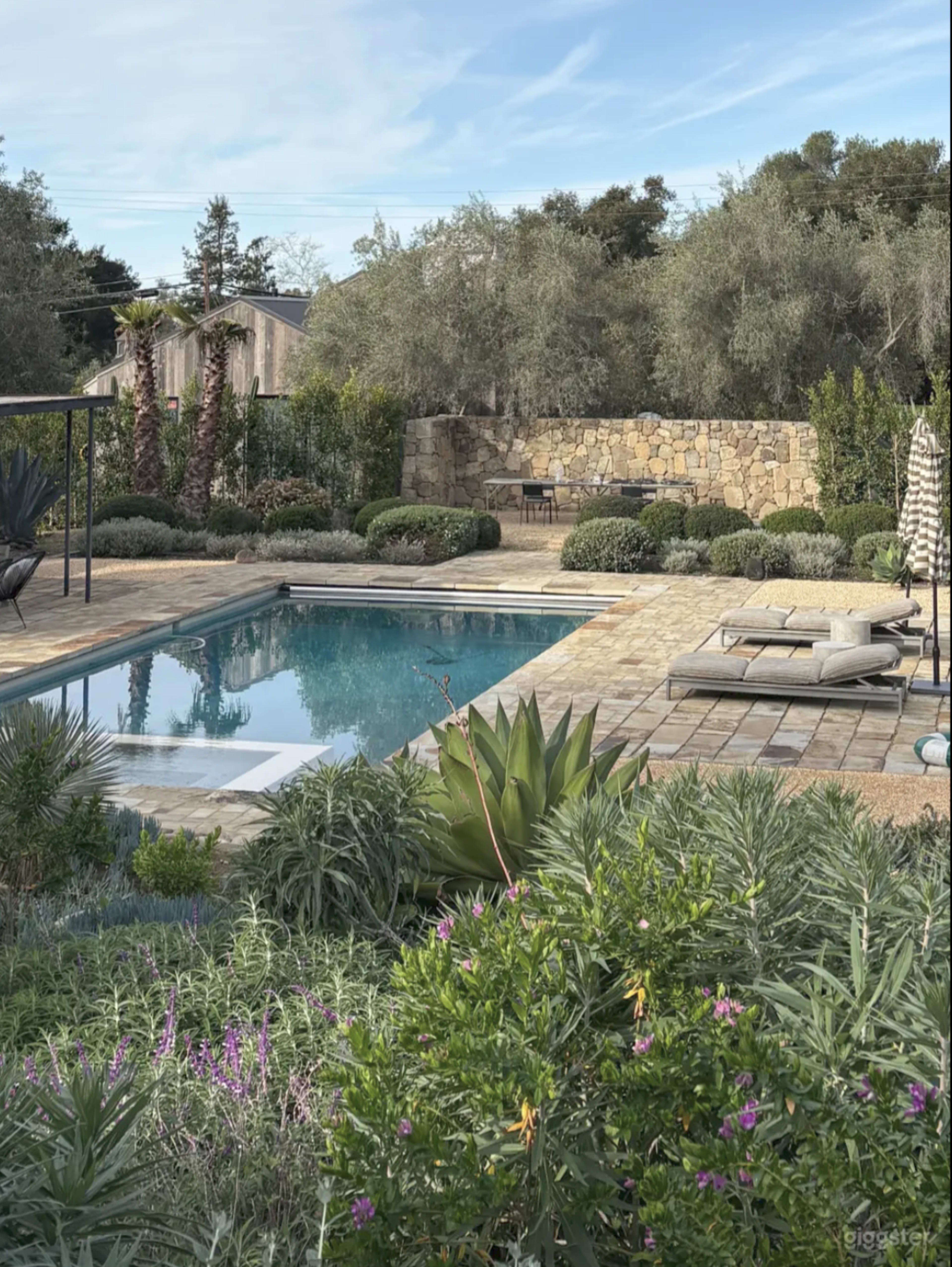 The image shows a landscaped garden with a swimming pool, surrounded by greenery and stone features, under a clear blue sky.