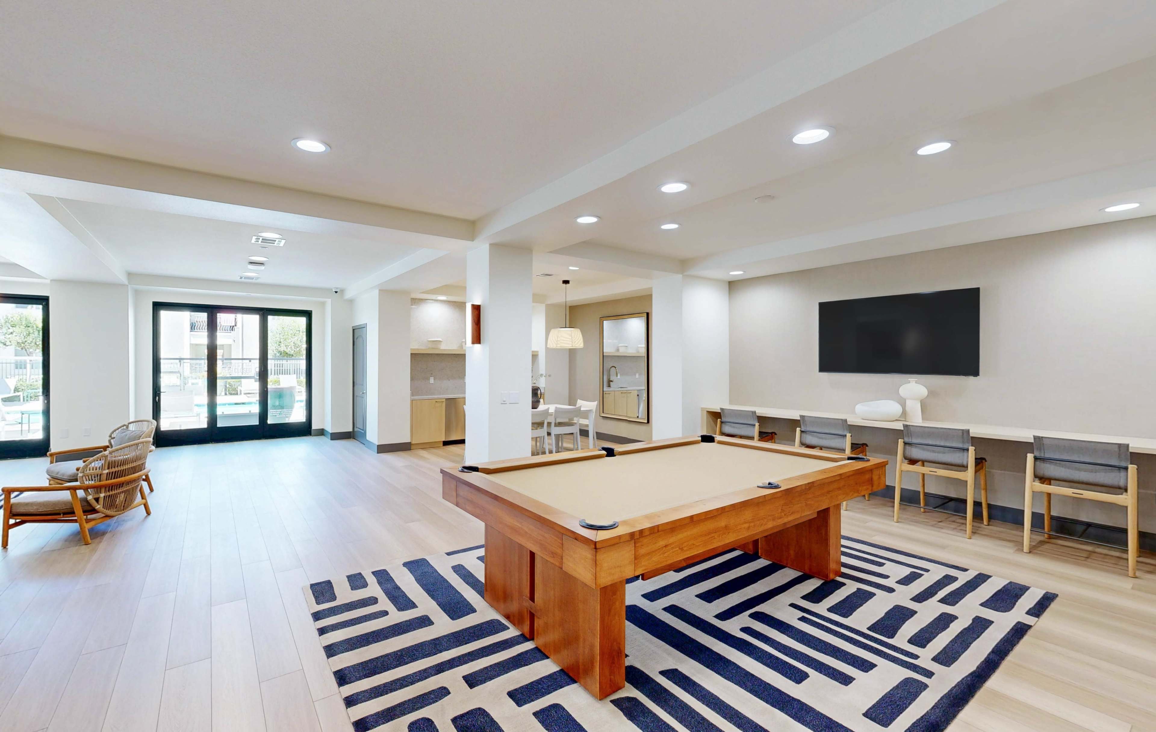 The image shows a spacious indoor recreational area featuring a pool table, a seating area, and a kitchenette, with large windows overlooking an outdoor space.