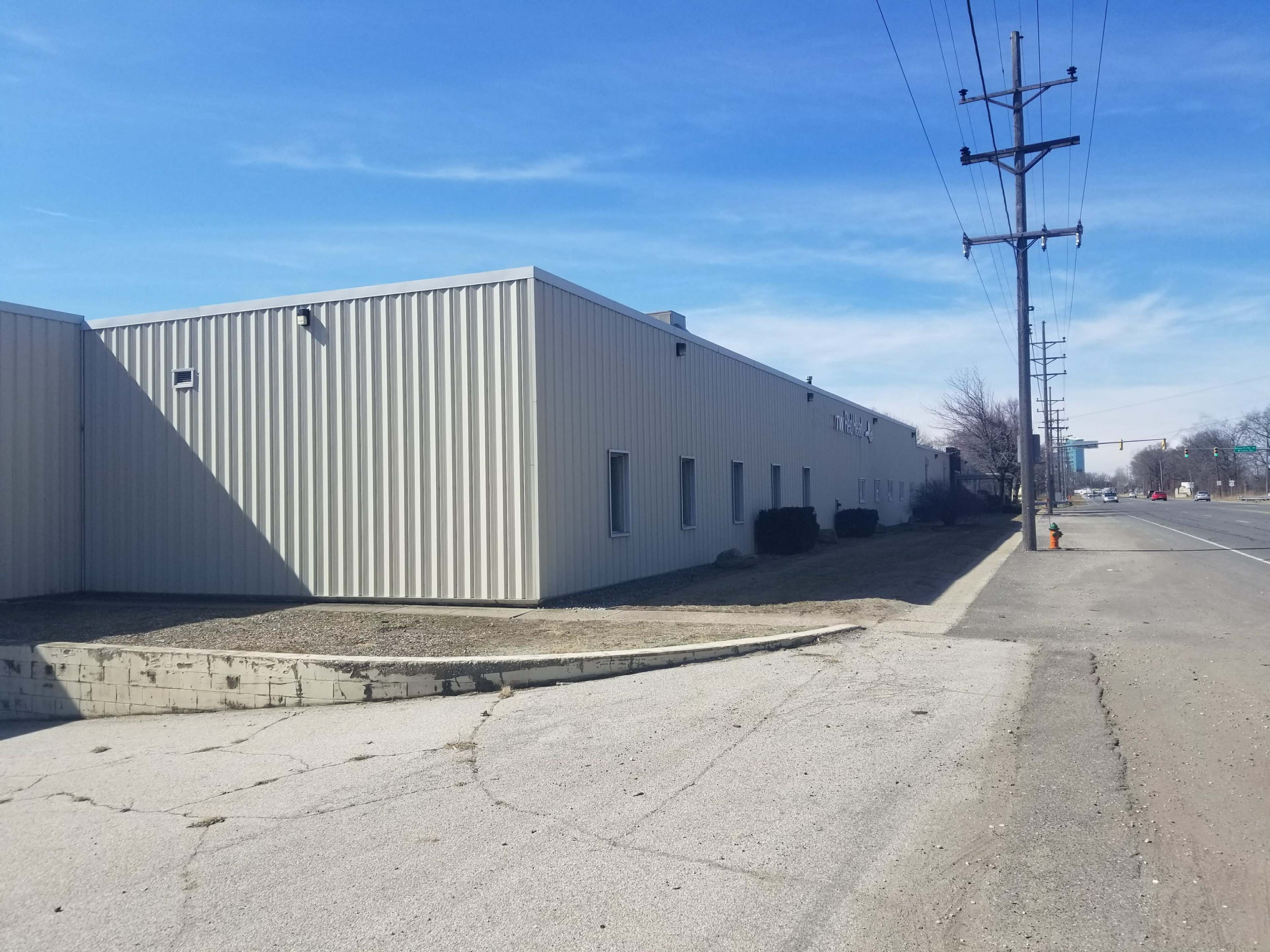 A light-colored metal building with a gravel area in front, located beside a paved road and power lines under a clear blue sky.