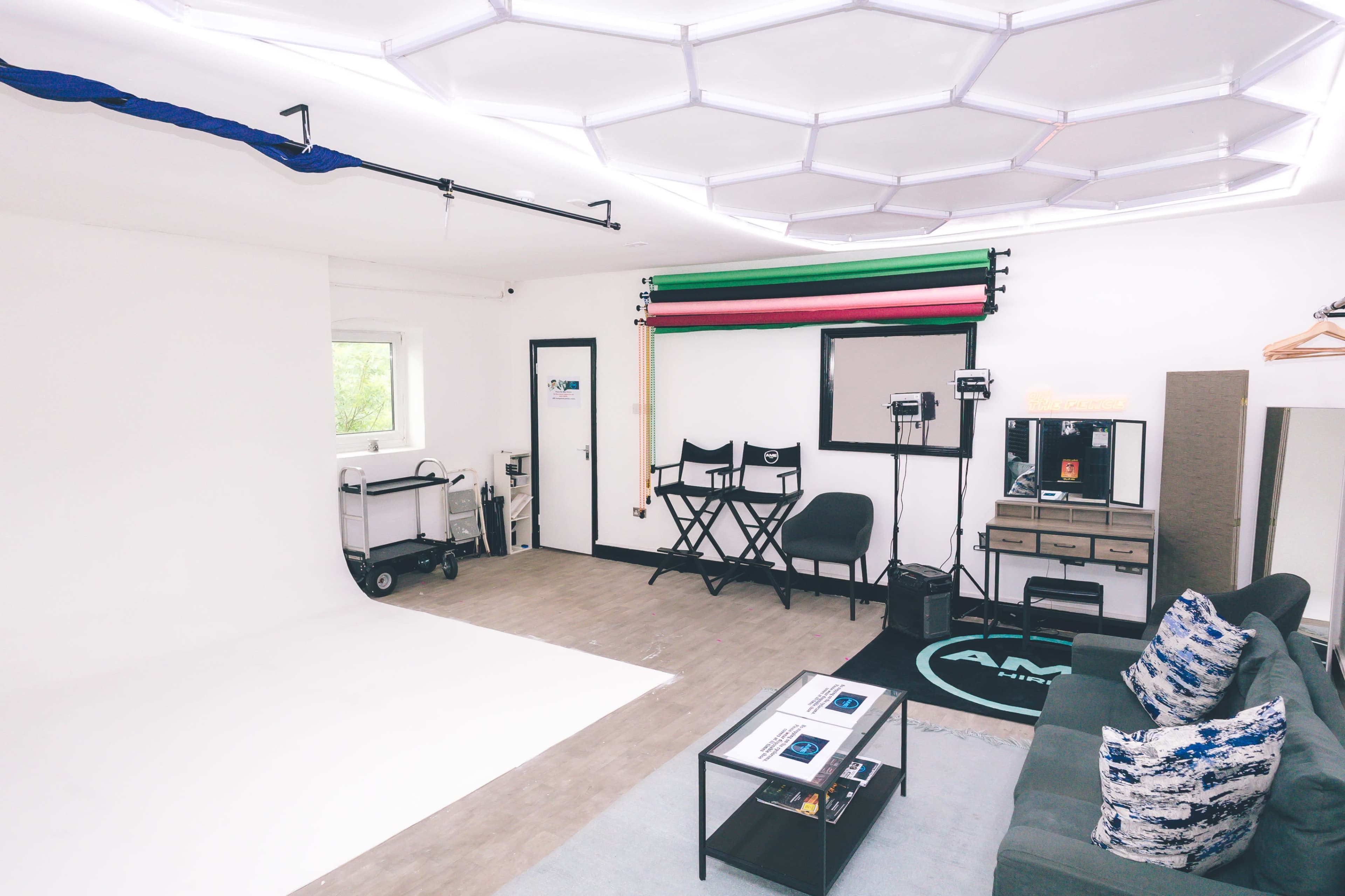 The image shows a well-lit photography studio featuring a seamless backdrop, makeup station, director's chairs, and equipment stands against a white wall.