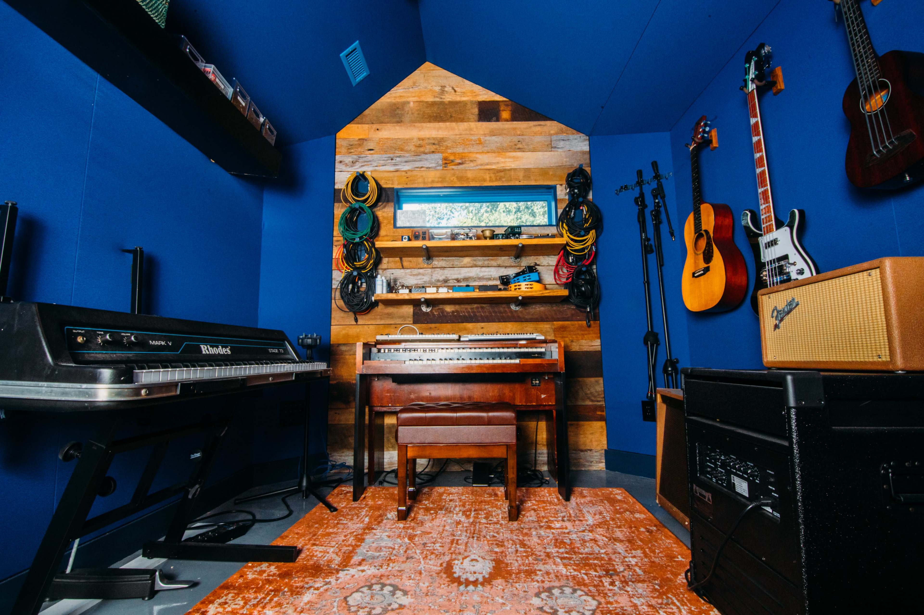 A music studio with a wooden desk holding a keyboard, flanked by various musical instruments and a colorful rug on the floor.