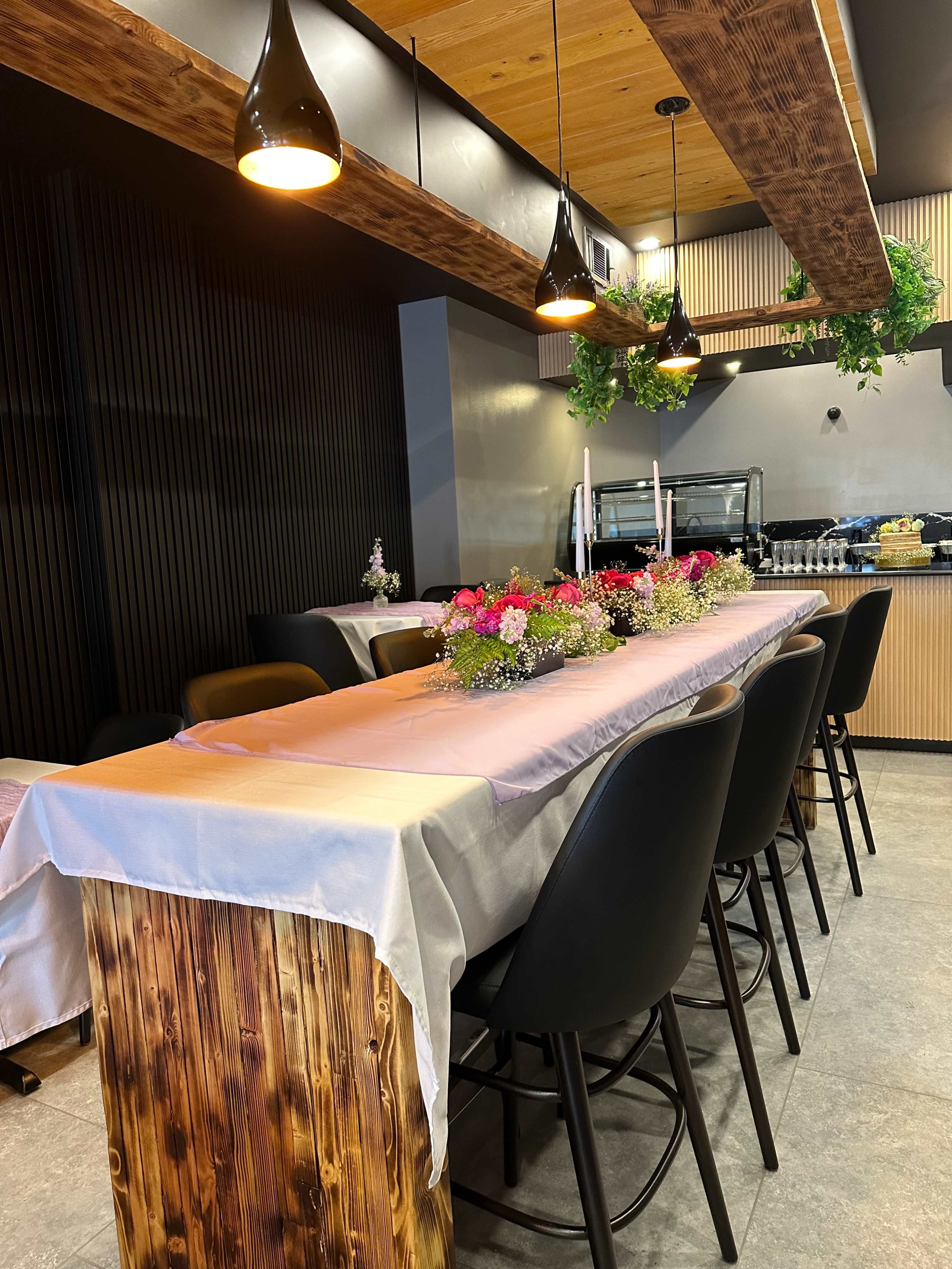 A long table is set with floral arrangements in a modern dining space featuring wooden accents and pendant lights.