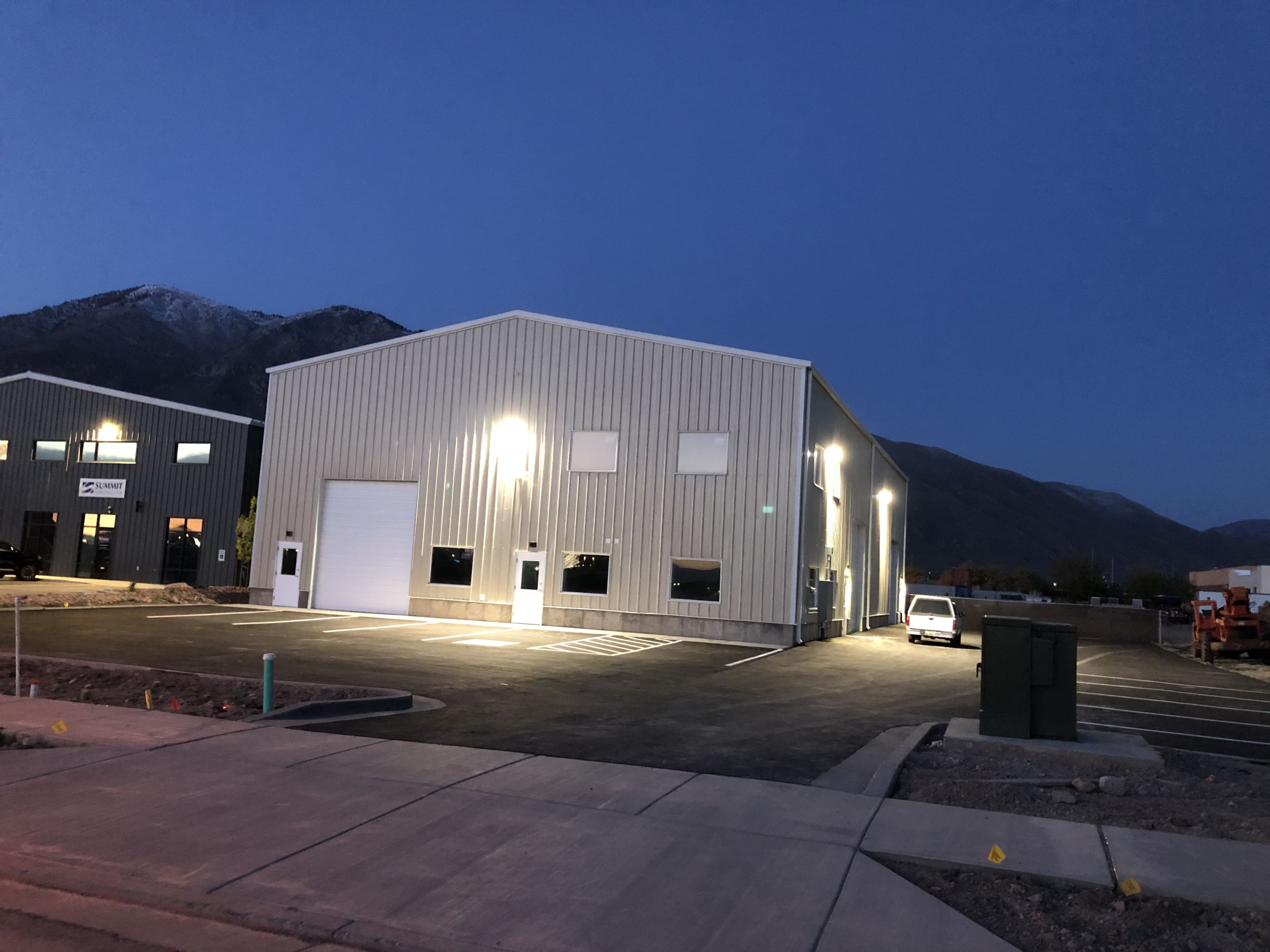 A large metal building with illuminated exterior lights stands in a parking lot near a mountain at dusk.