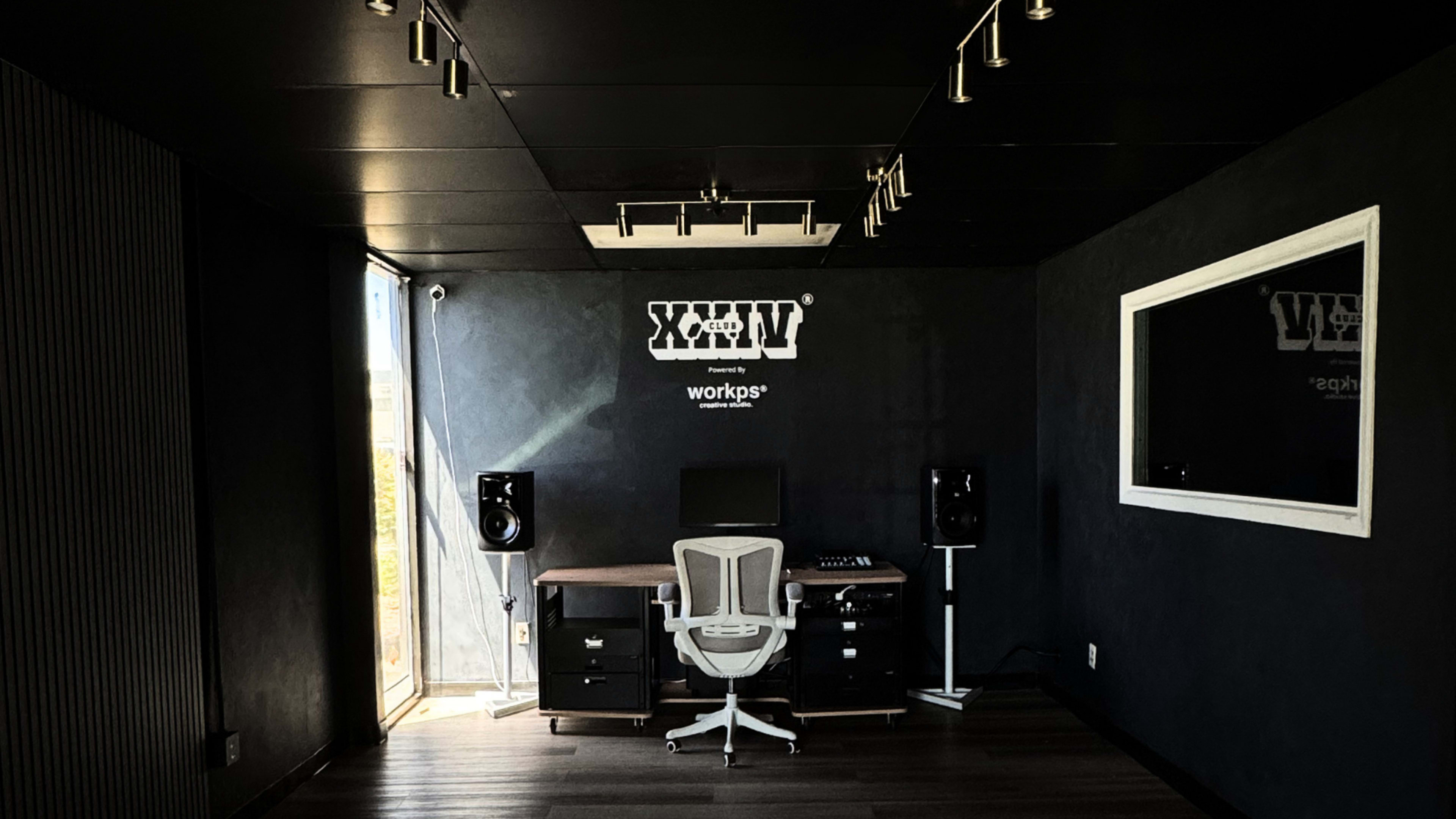The image shows a minimalist workroom featuring a desk with a computer setup, two speakers, and a large window allowing natural light.